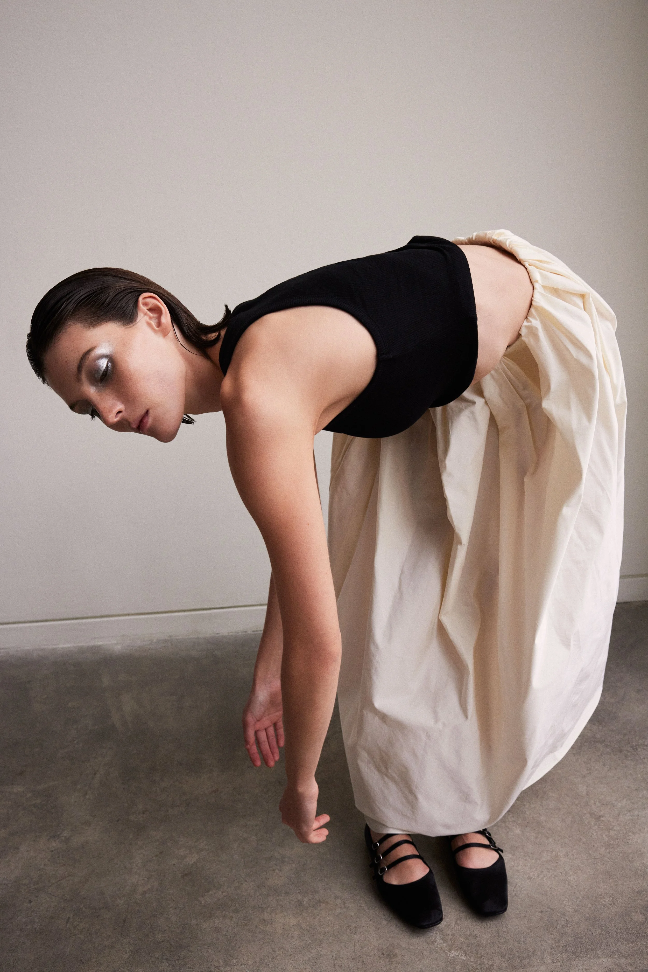 A woman with short dark hair and silver eyeshadow is bent over, looking down, wearing a black sleeveless top, a long cream-colored skirt, and black shoes.