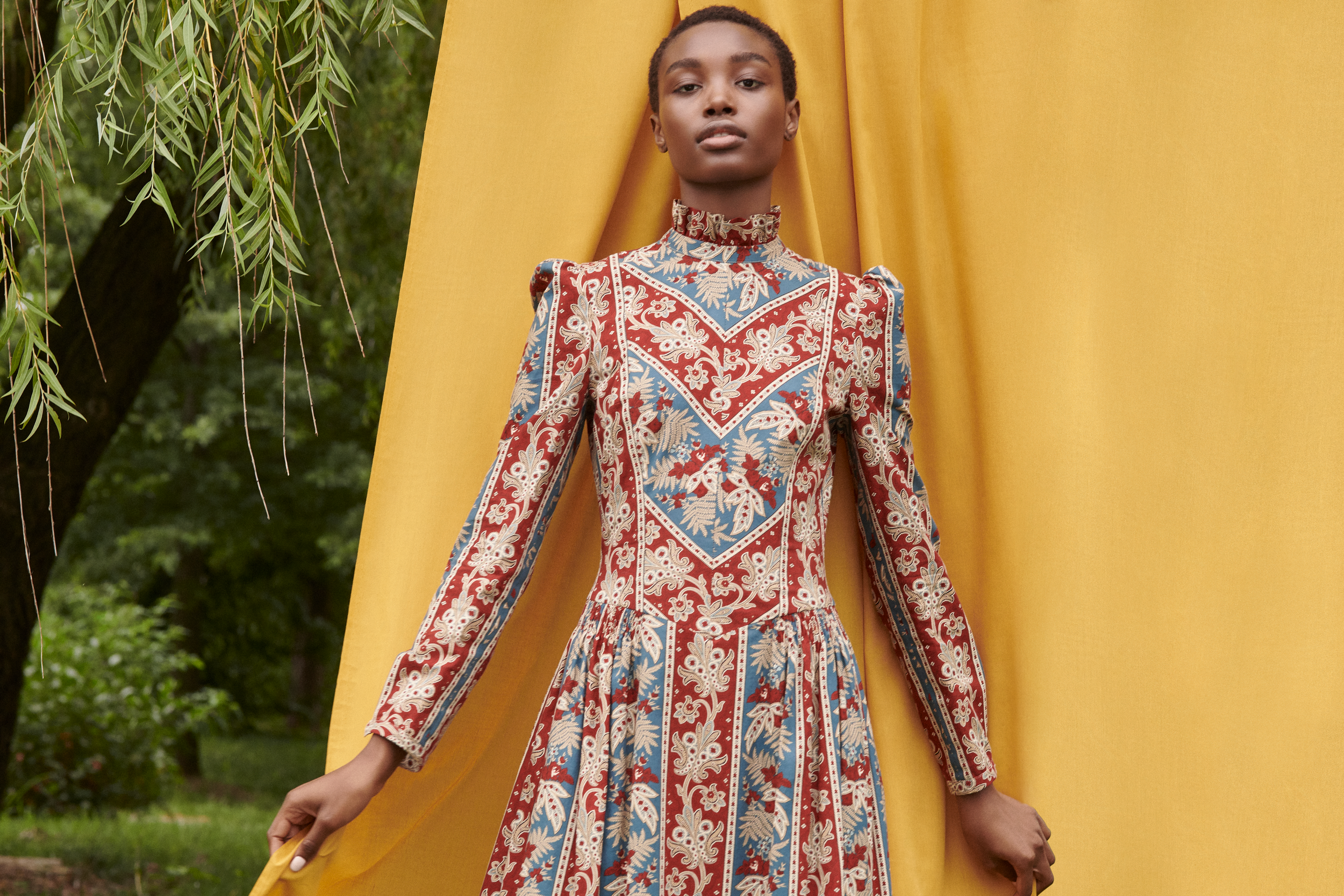 A woman wearing a long-sleeved, high-neck dress with an intricate red, blue, and beige floral pattern, standing outdoors with a yellow curtain hanging behind her.