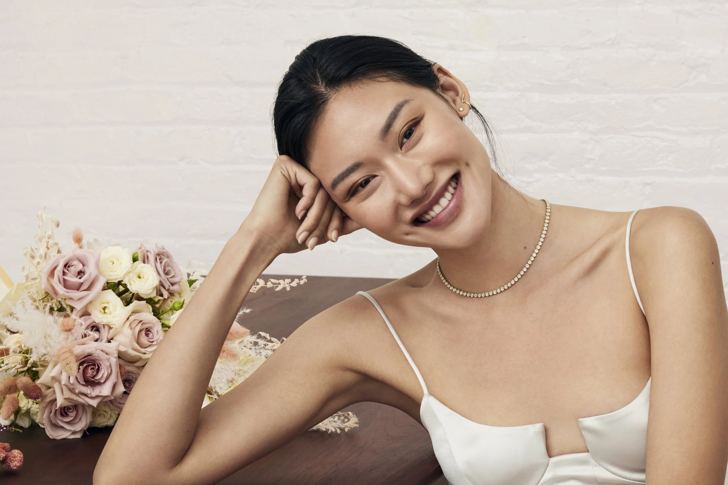 A young woman with short black hair, smiling and resting her head on her hand, sitting at a table with a bouquet of pink and white roses beside her, wearing a white spaghetti strap top and a pearl necklace, against a white brick wall background.