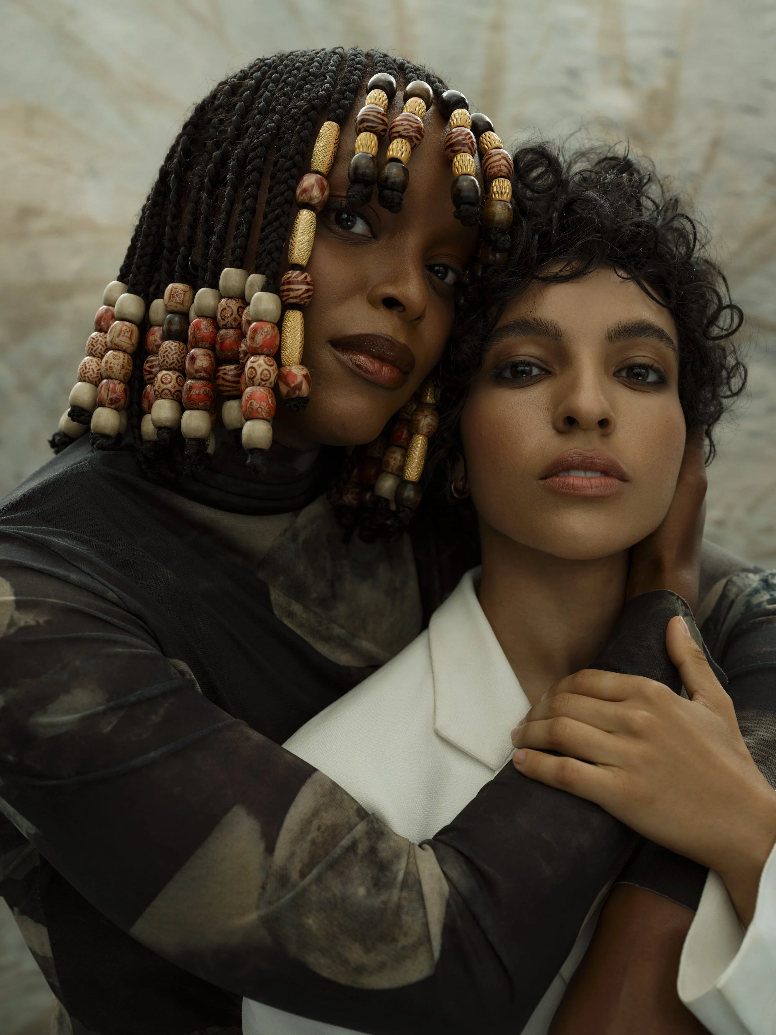 Two women embrace closely, one with dark skin and colorful beaded braids, the other with light skin and curly short hair, both looking at the camera against a marble background.