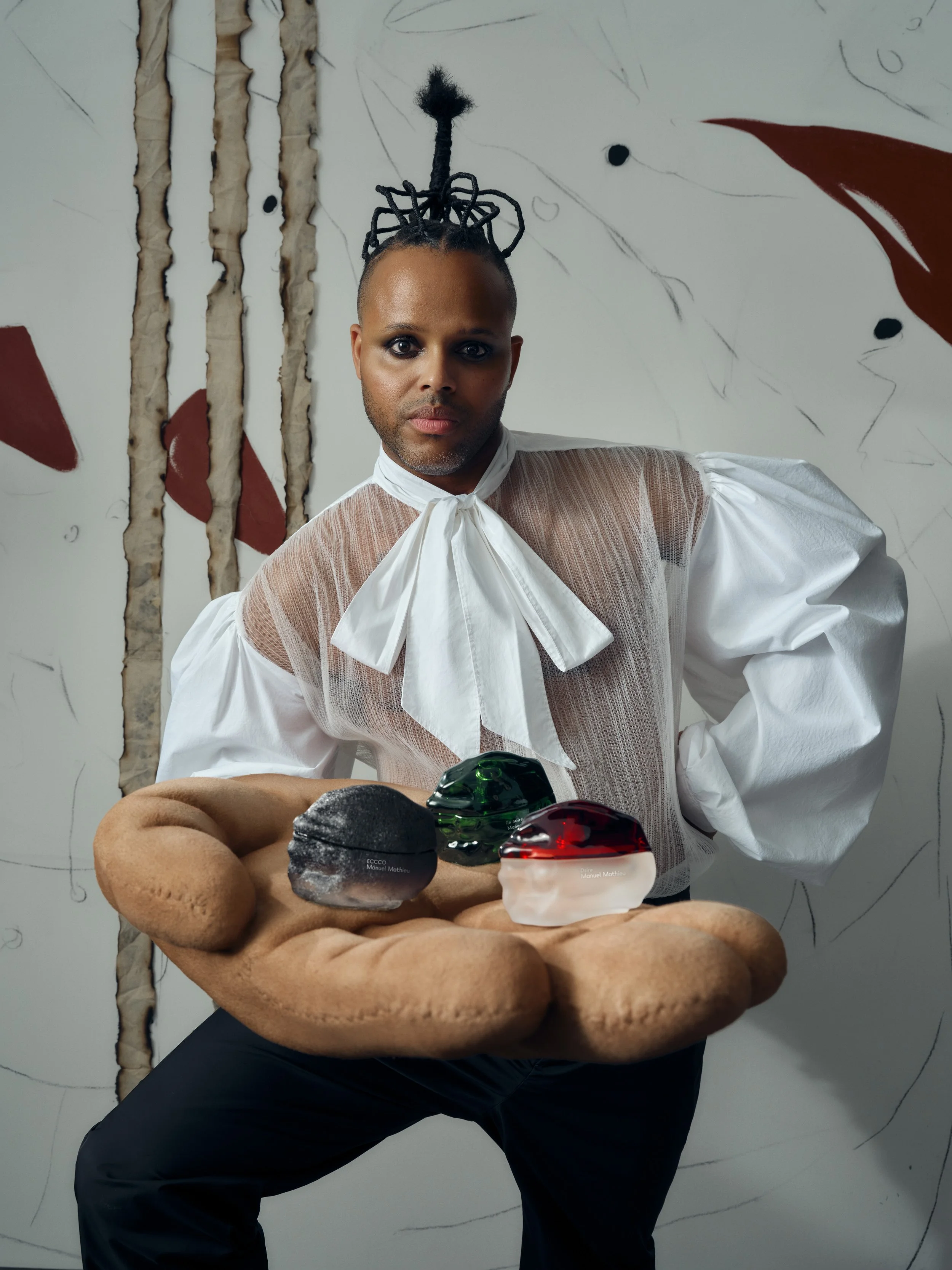 Person with styled hair and dressed in a sheer white blouse with a large bow, holding a large plush hand displaying three small decorative objects, against an abstract background.