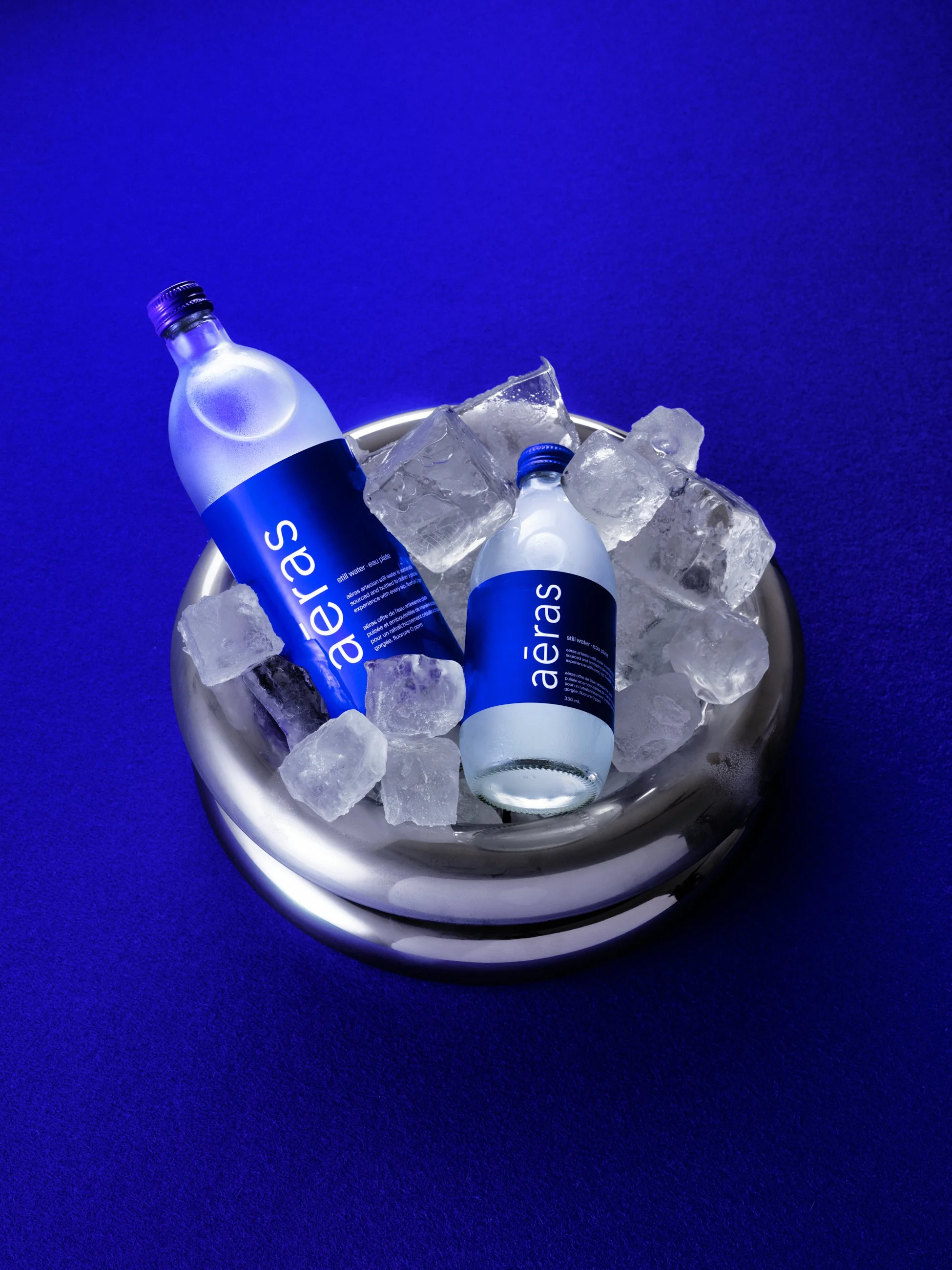 Two bottles of Aéreas water in a silver bowl filled with ice cubes, set on a blue surface with a dark blue background.