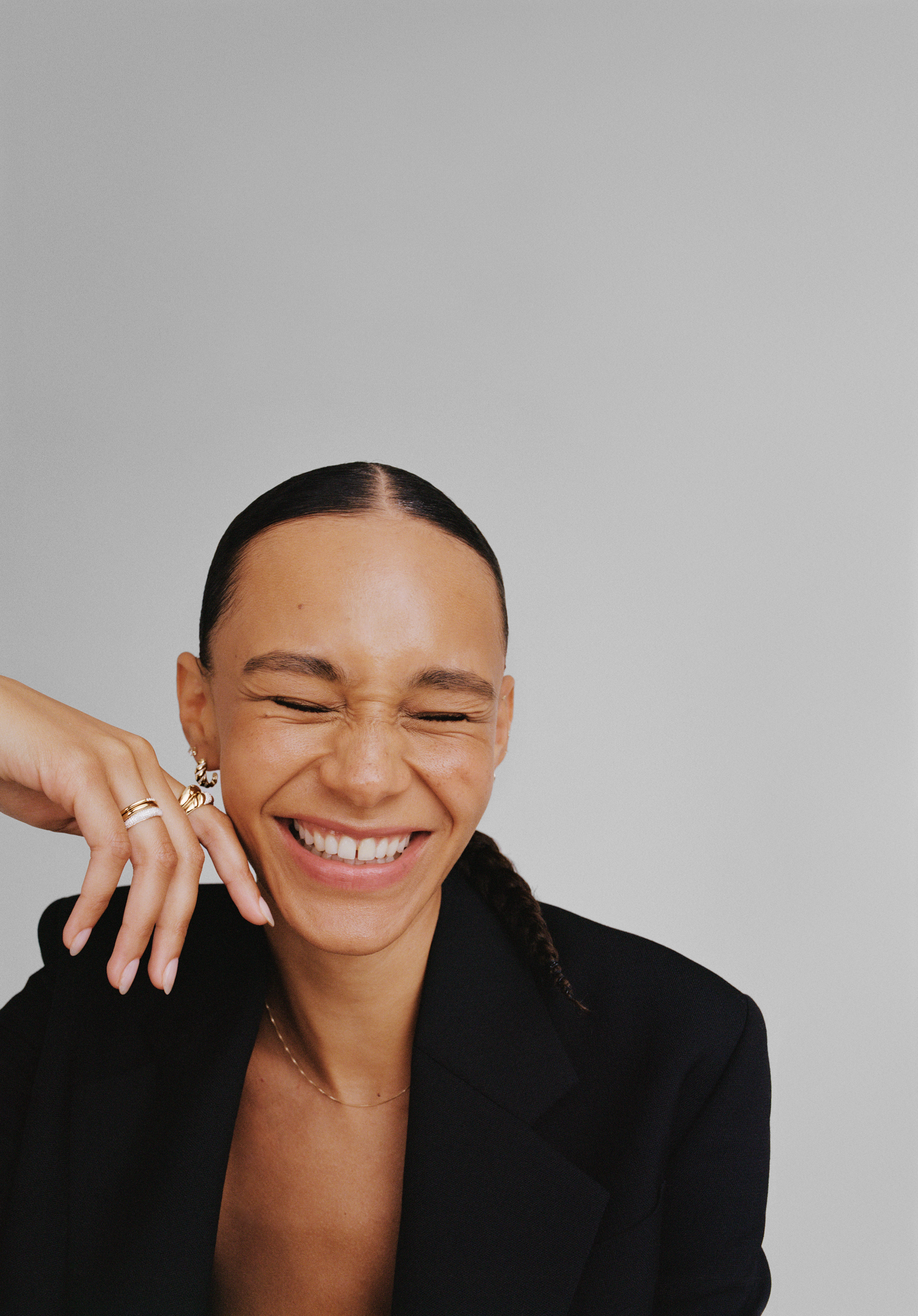 A woman with dark hair styled in a braid, laughing with eyes closed, wearing gold jewelry and a black blazer, against a plain light gray background.