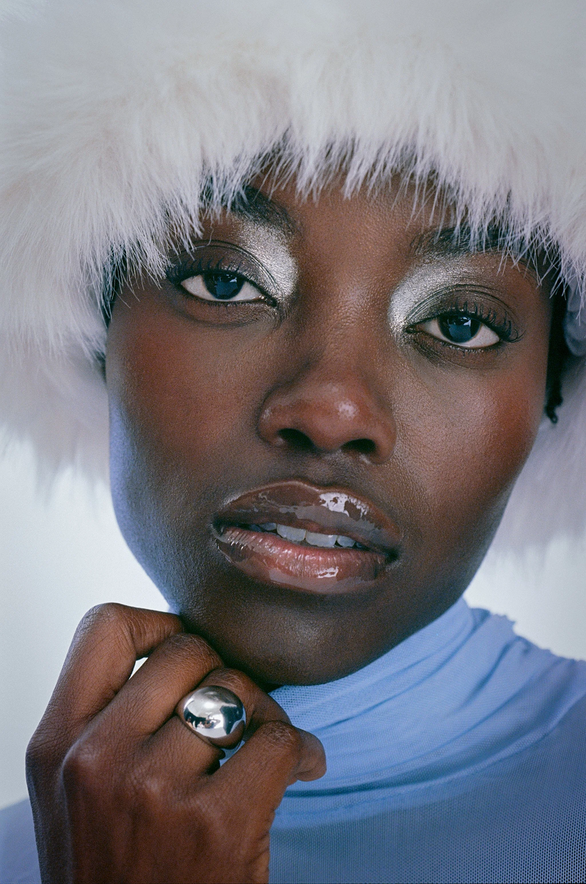 Close-up of a woman wearing a white furry hat with silver eye makeup, glossy lips, and a reflective silver ring on her finger, with a light blue top.