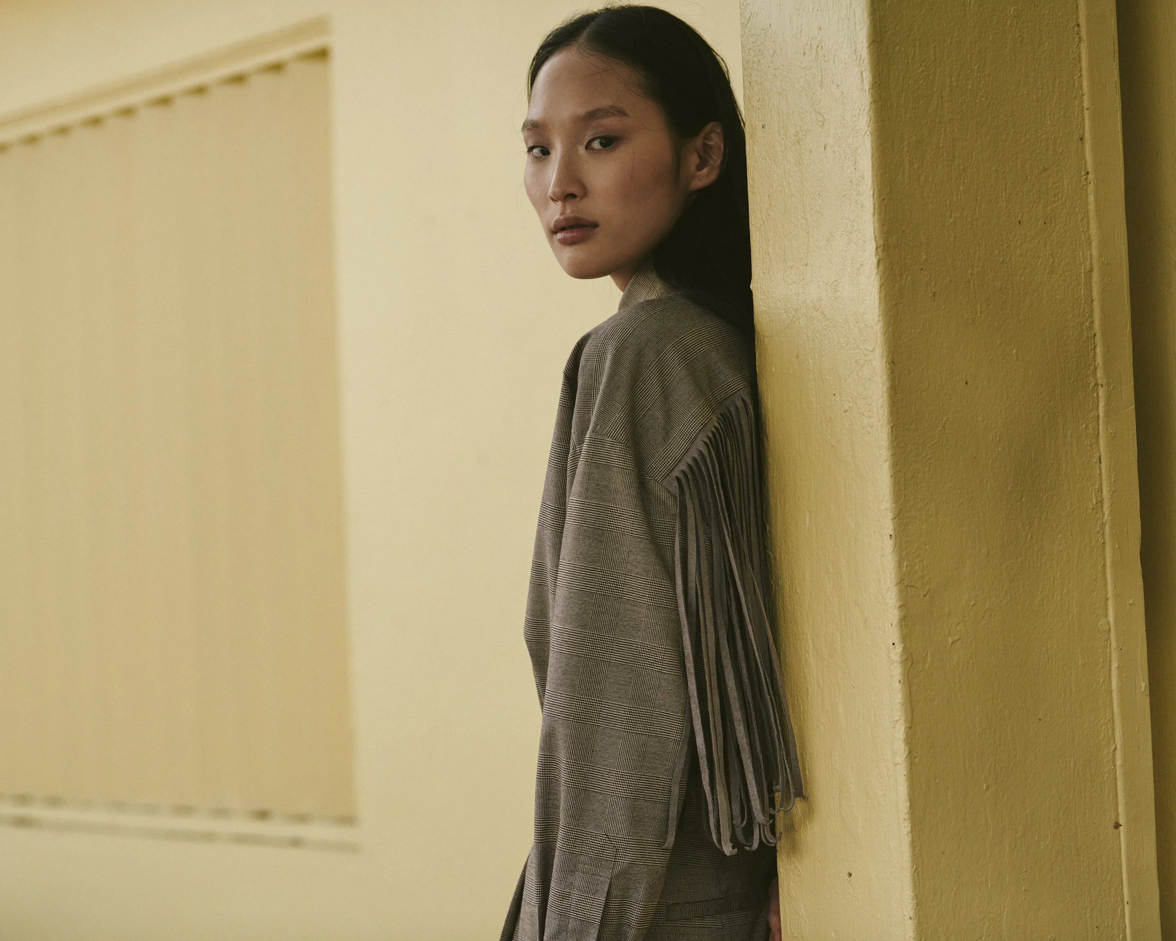 A woman with long dark hair leaning against a yellow wall, wearing a plaid blazer with fringe details on the sleeves, looking at the camera with a neutral expression.