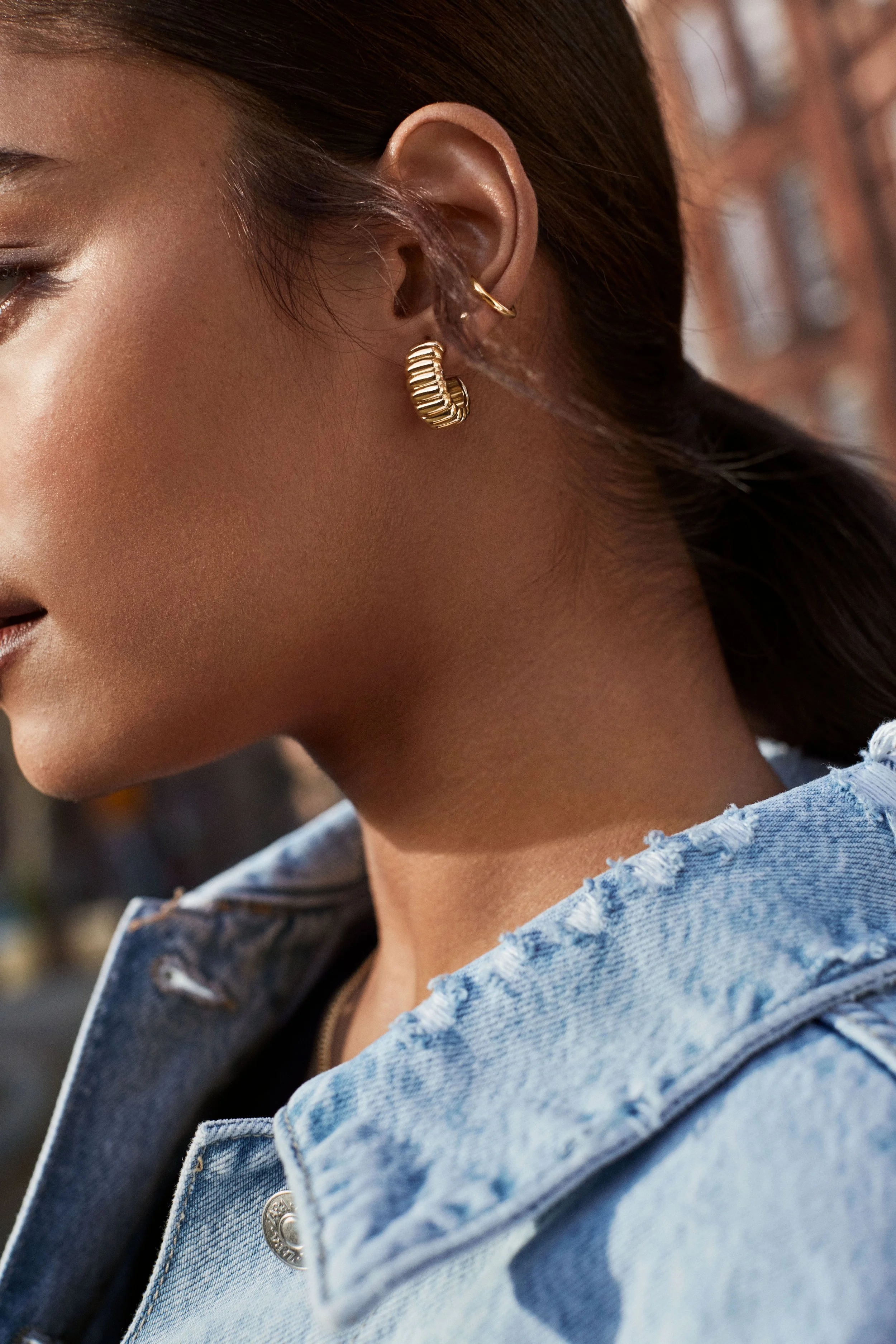 Close-up of a woman wearing gold hoop earrings and a distressed denim jacket, with sunlight highlighting her face and neck, against a blurred urban background.