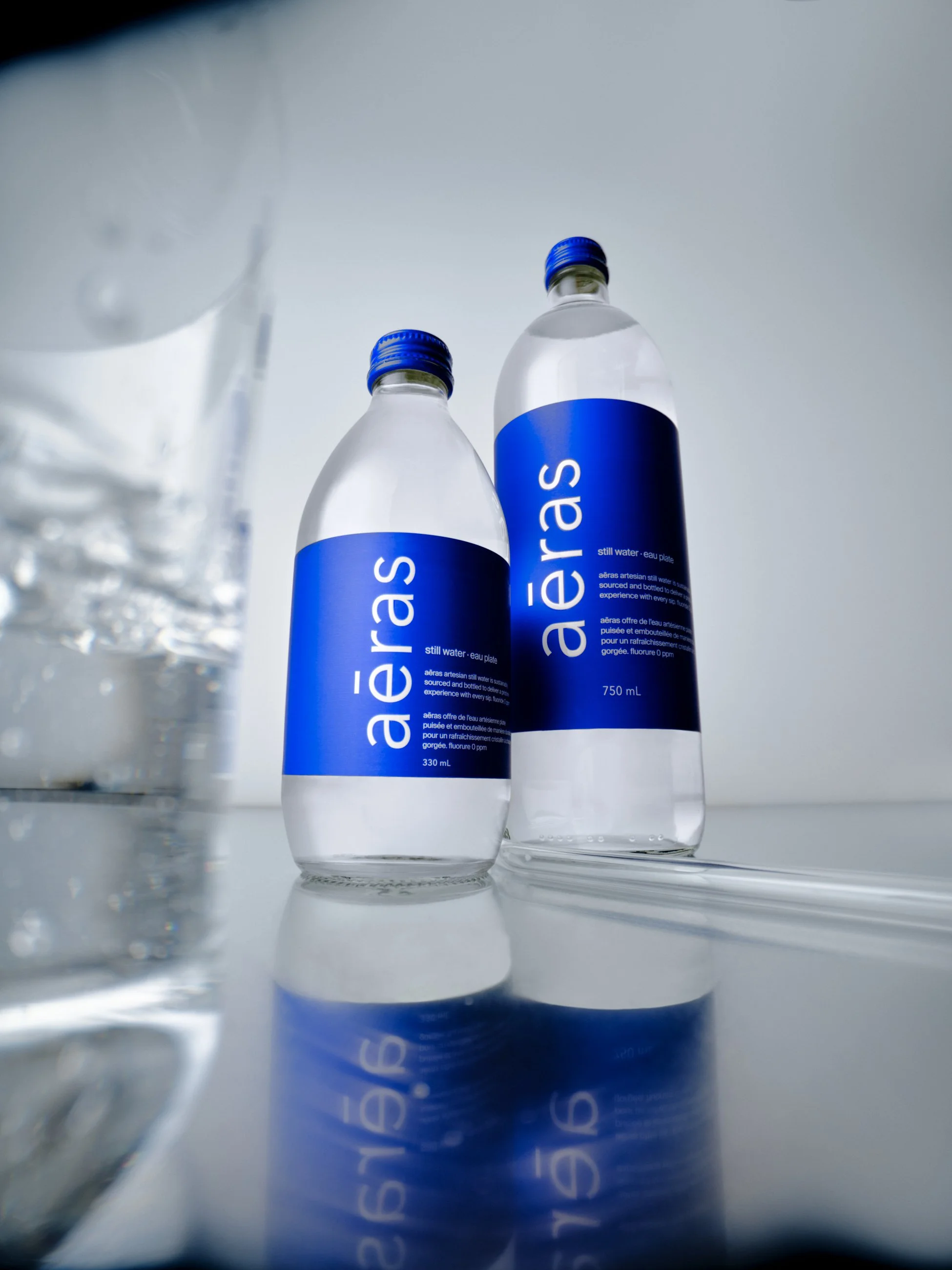 Two bottles of still water with blue labels and caps, placed next to each other on a reflective surface, with a glass of water partially visible on the left.