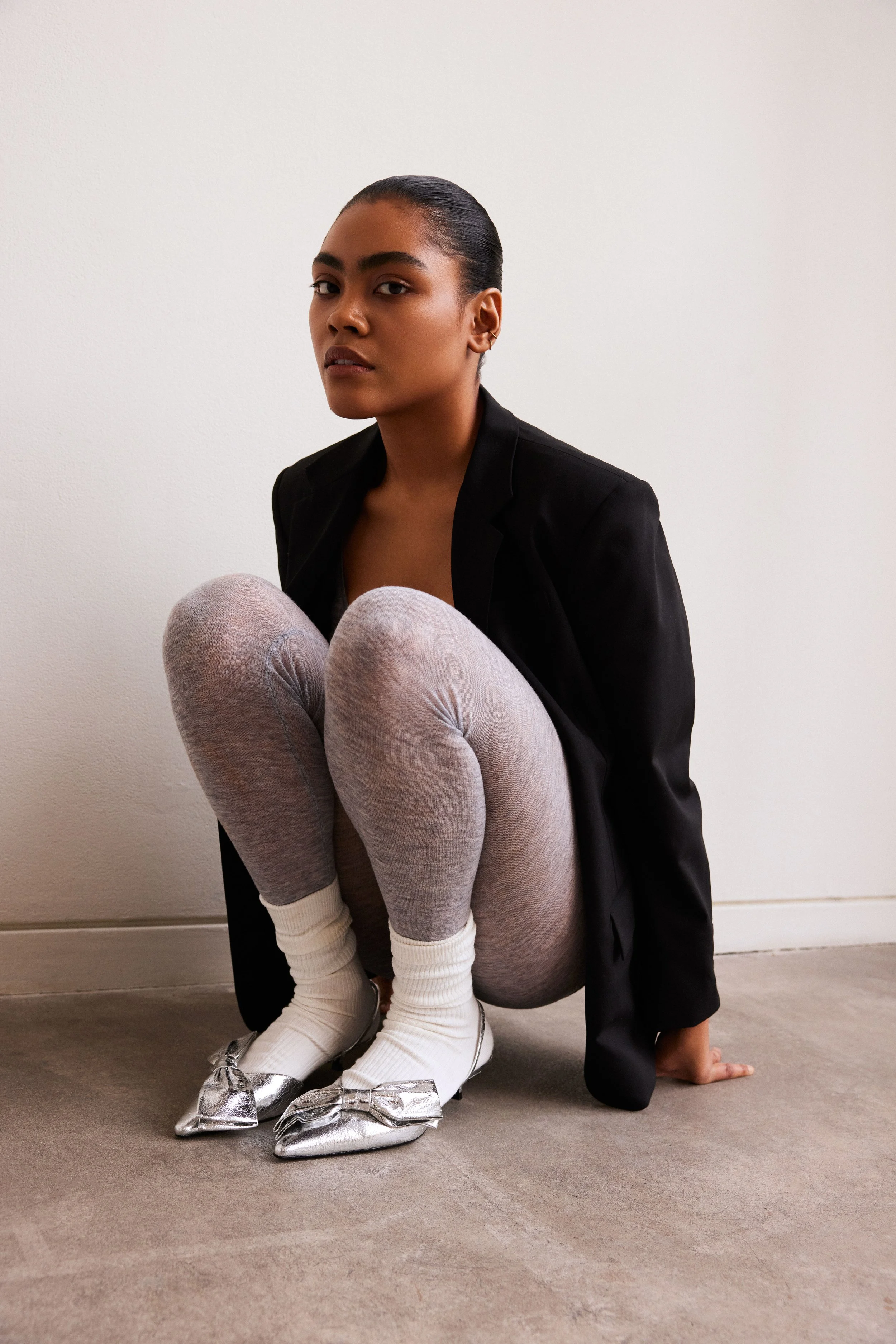 A woman with dark hair pulled back, dressed in a black blazer, crouched on the floor against a plain white wall. She is wearing light gray leggings and silver pointed-toe flats with bows, along with white socks.