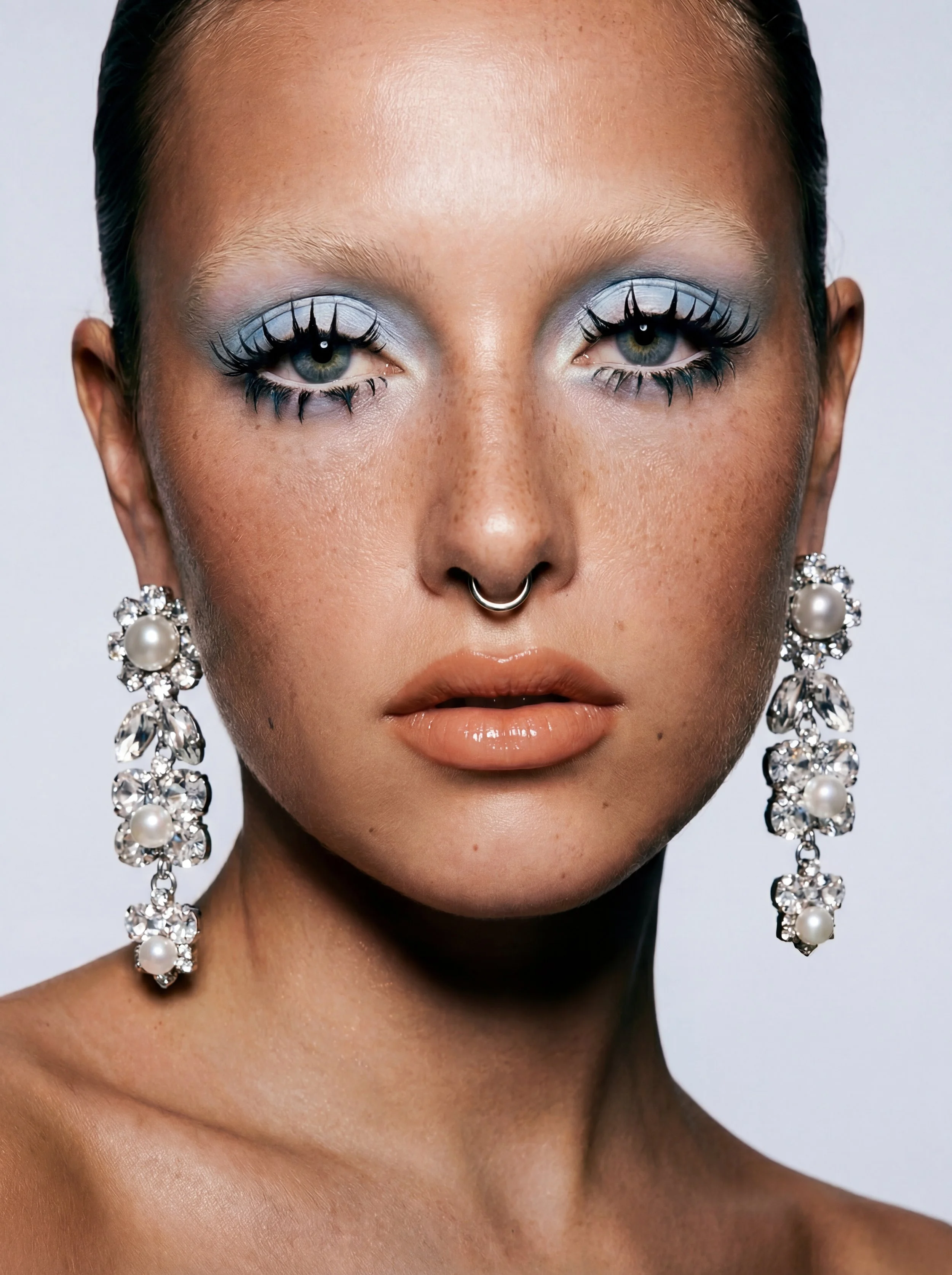 Close-up portrait of a woman with makeup, wearing large chandelier earrings with pearls and rhinestones, a septum piercing, and slicked-back dark hair.