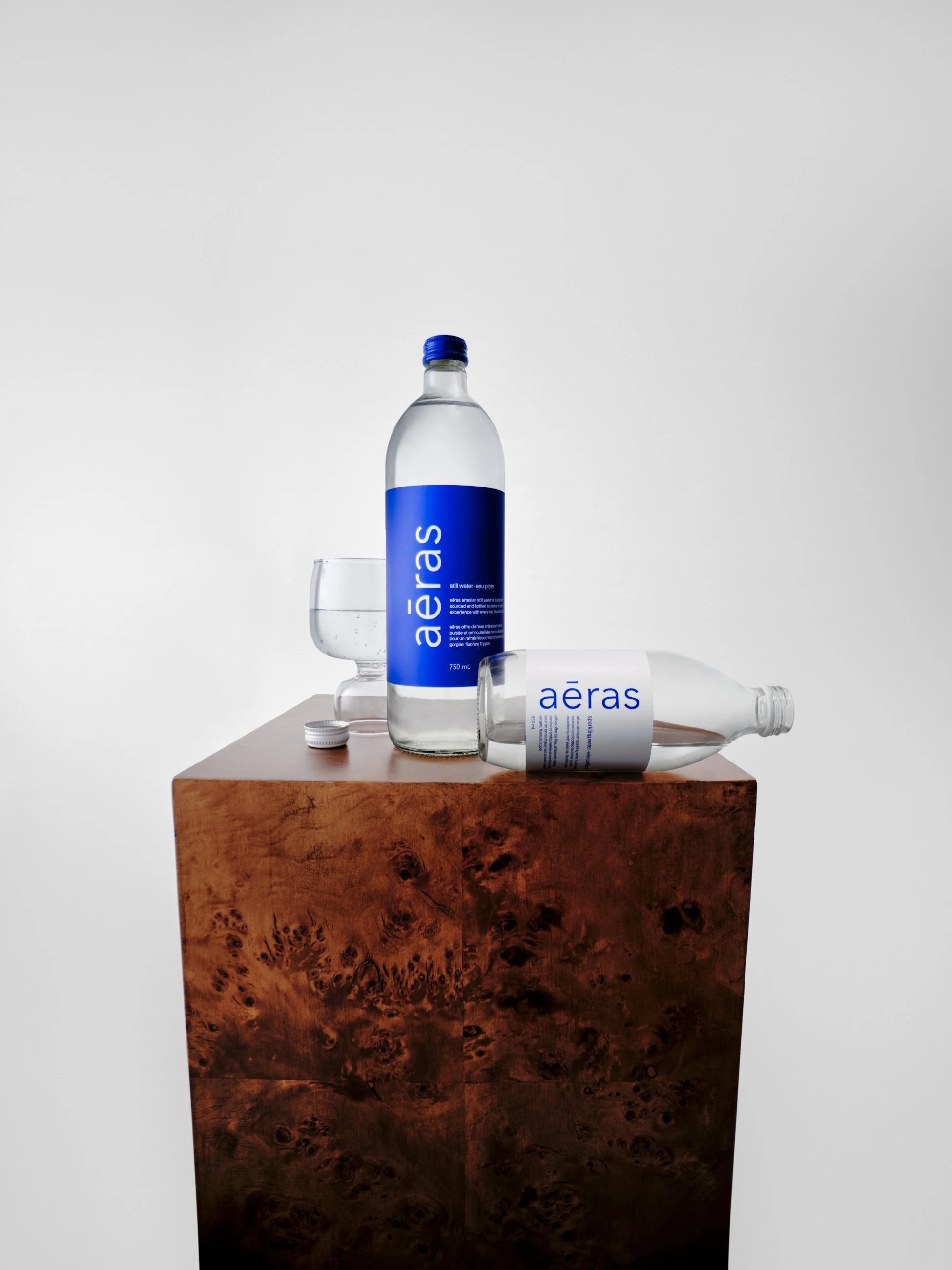 A bottle of Aeras mineral water, a glass of water, and another bottle lying on its side on a wooden surface against a plain white background.