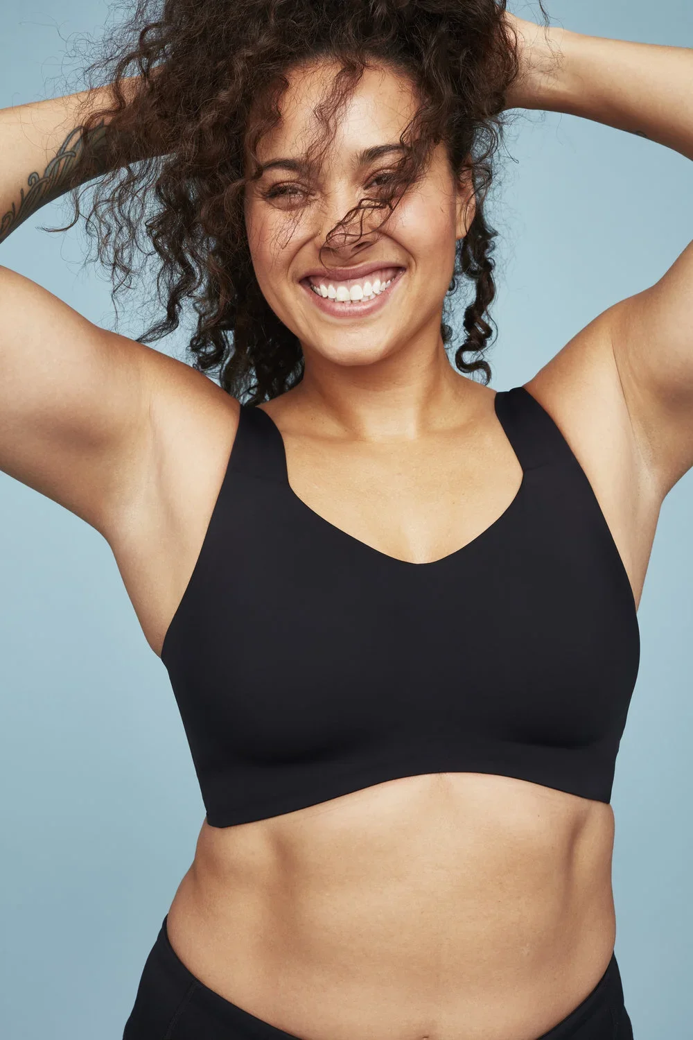 A smiling woman with curly hair wearing a black sports bra against a light blue background.