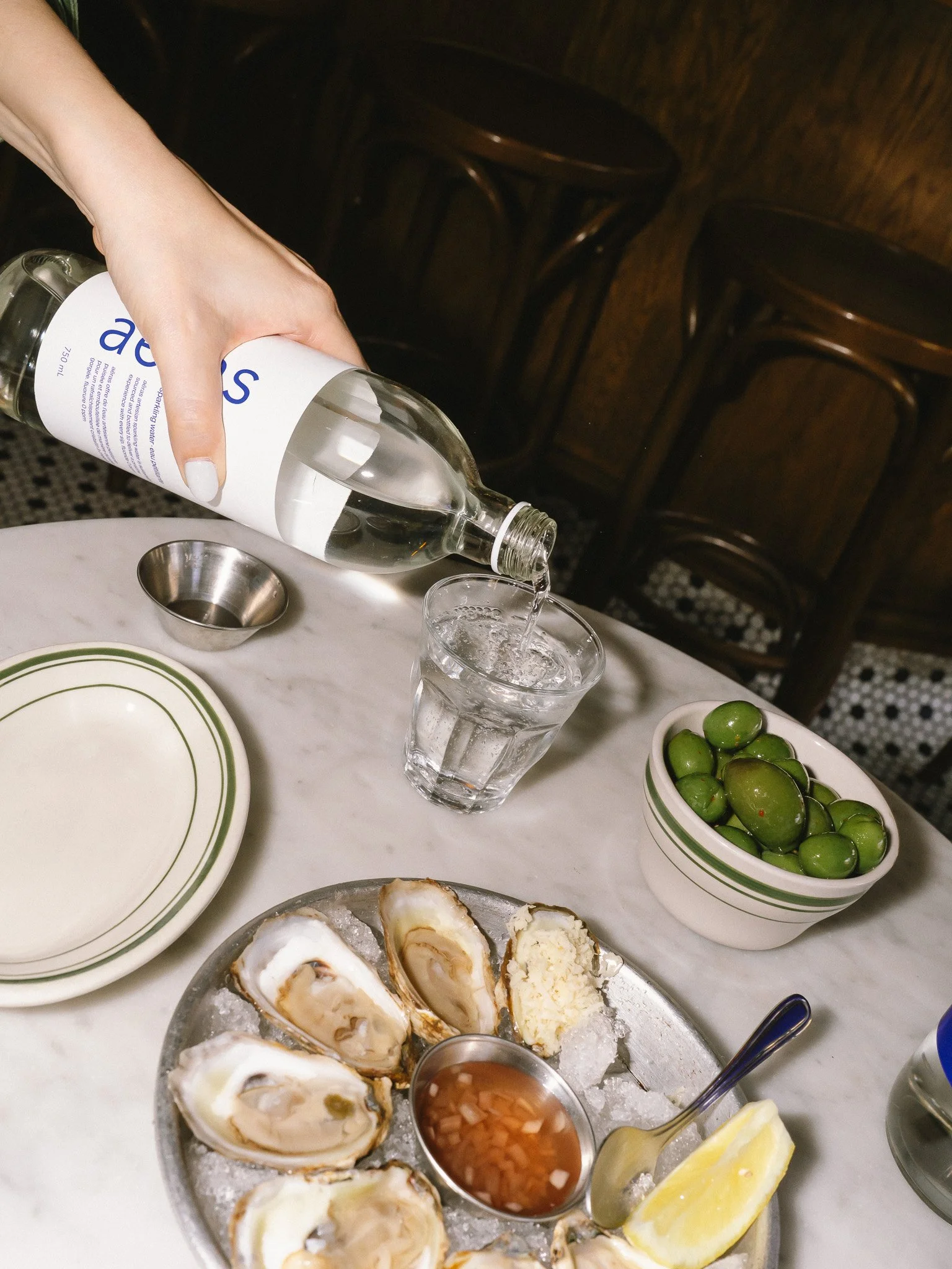 Person pouring water into a glass at a dining table with oysters on a plate, green olives in a bowl, a lemon wedge, and a small bowl of cocktail sauce.
