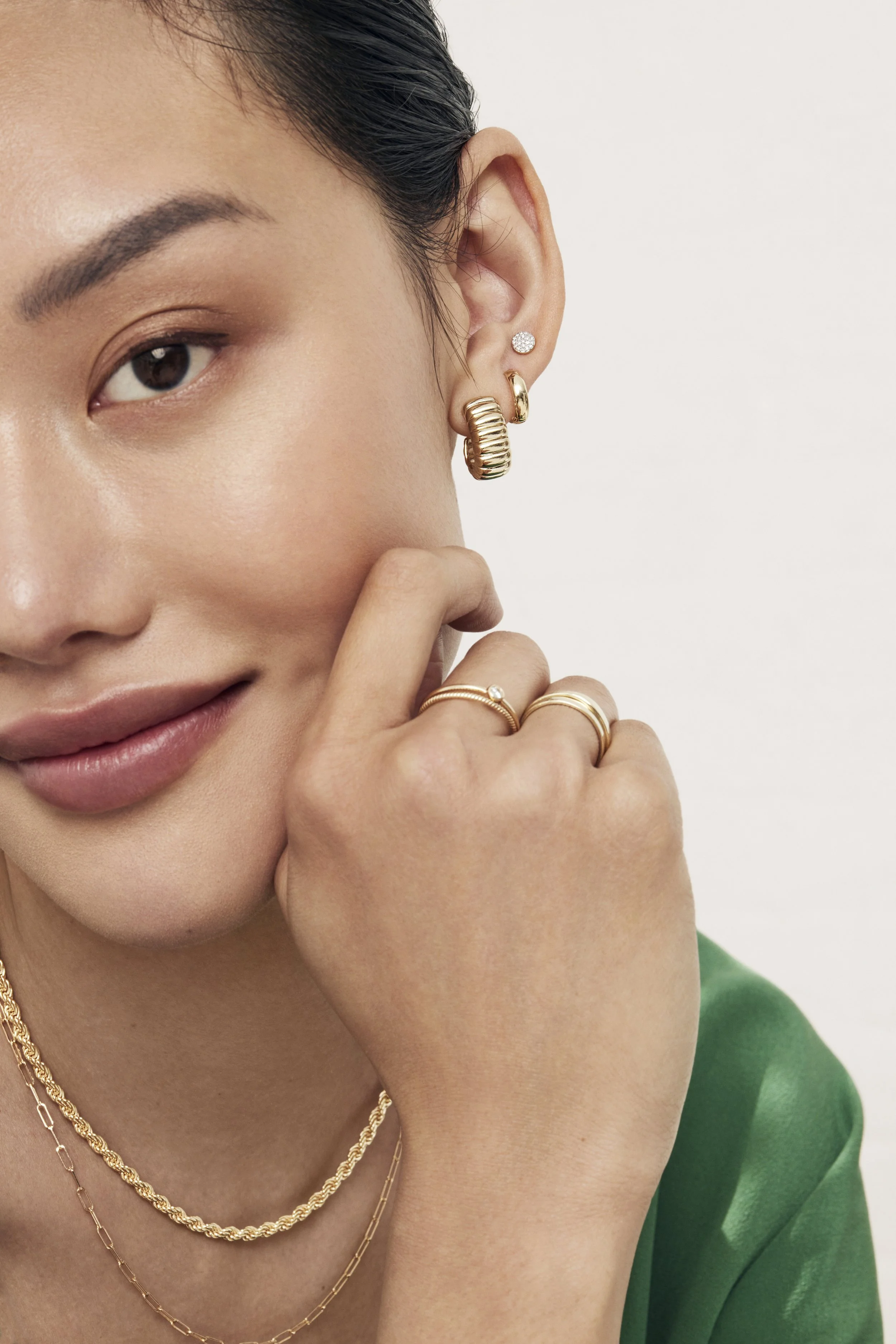 Close-up of a woman with dark hair and earrings, wearing layered gold necklaces and rings, dressed in a green top.