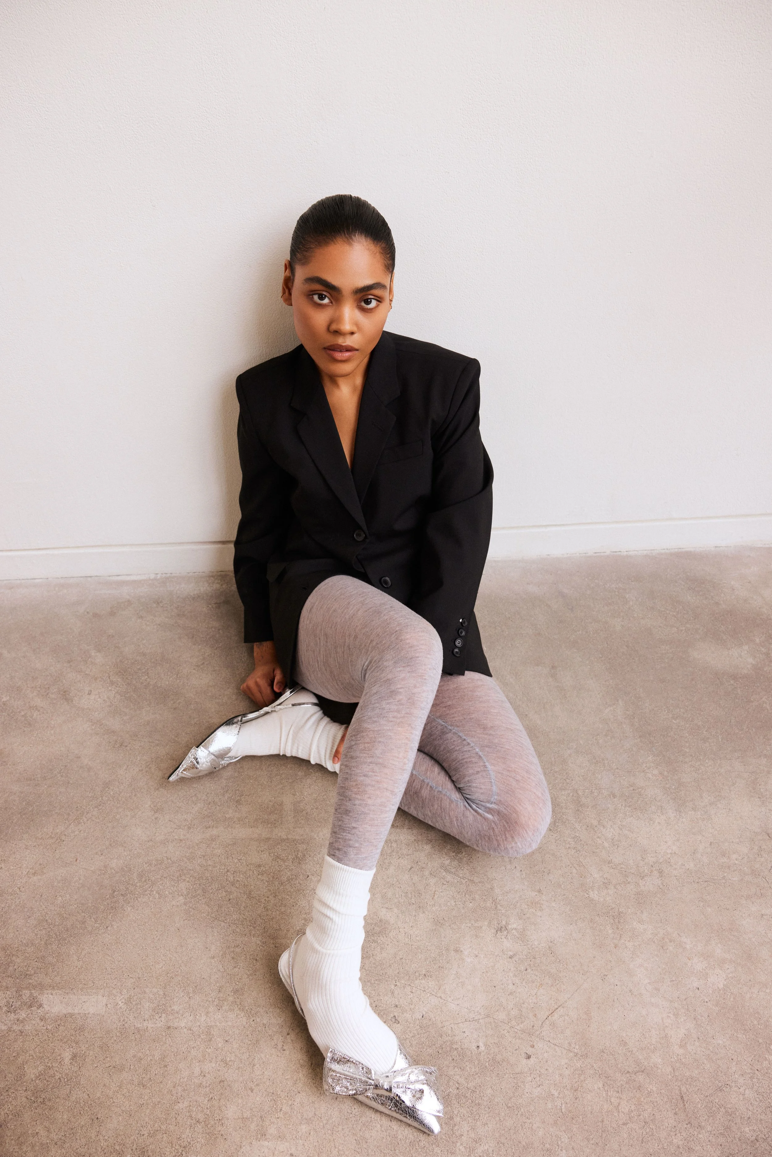 A young woman sitting on the floor against a plain white wall, wearing a black blazer, gray leggings, and silver metallic pointed-toe shoes with bows, with a neutral expression and dark hair in a neat bun.