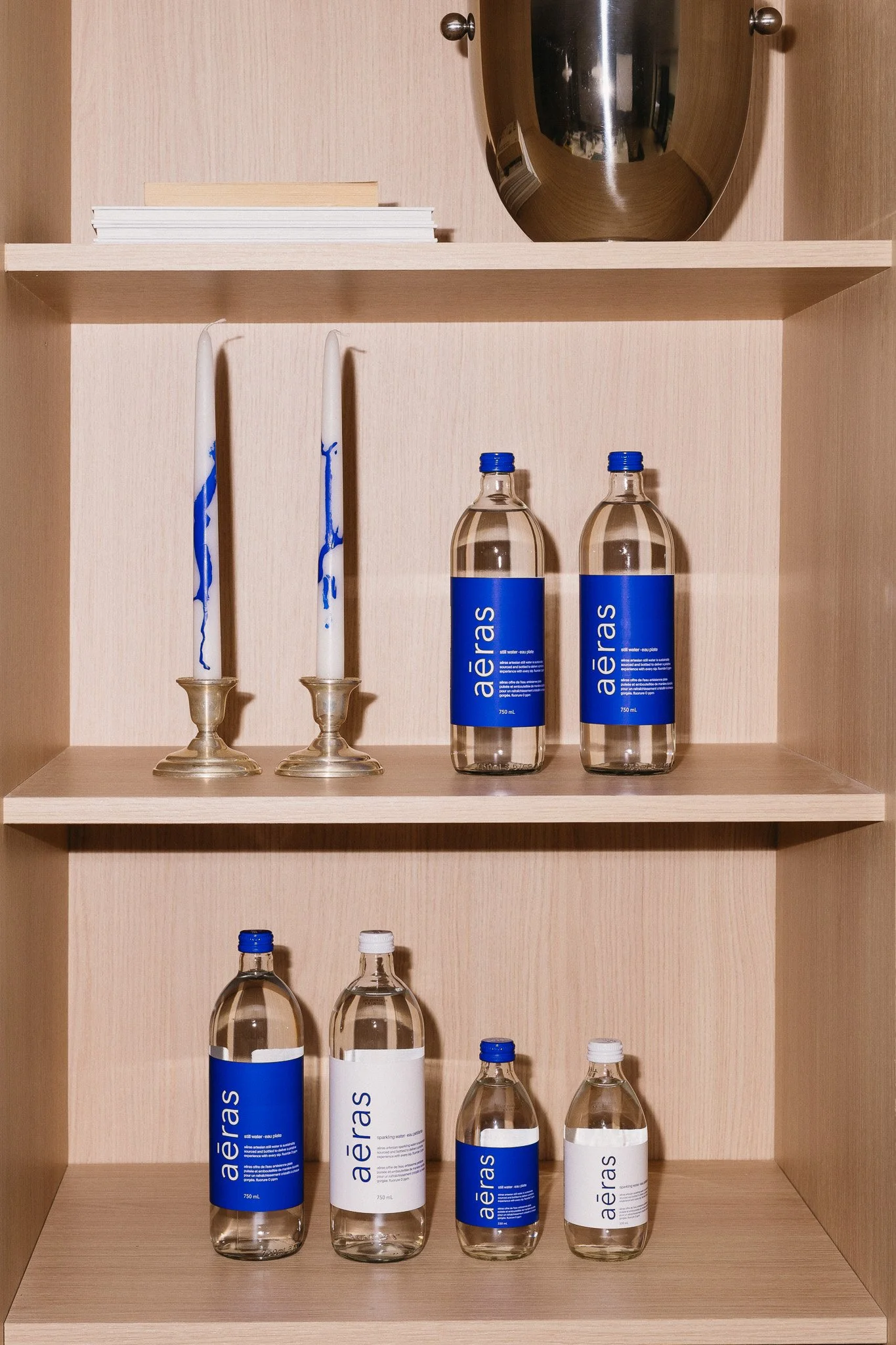 Decorative display of two white candles with blue wax drips, six bottles of water with blue and white labels, on wooden shelves against a beige wall.