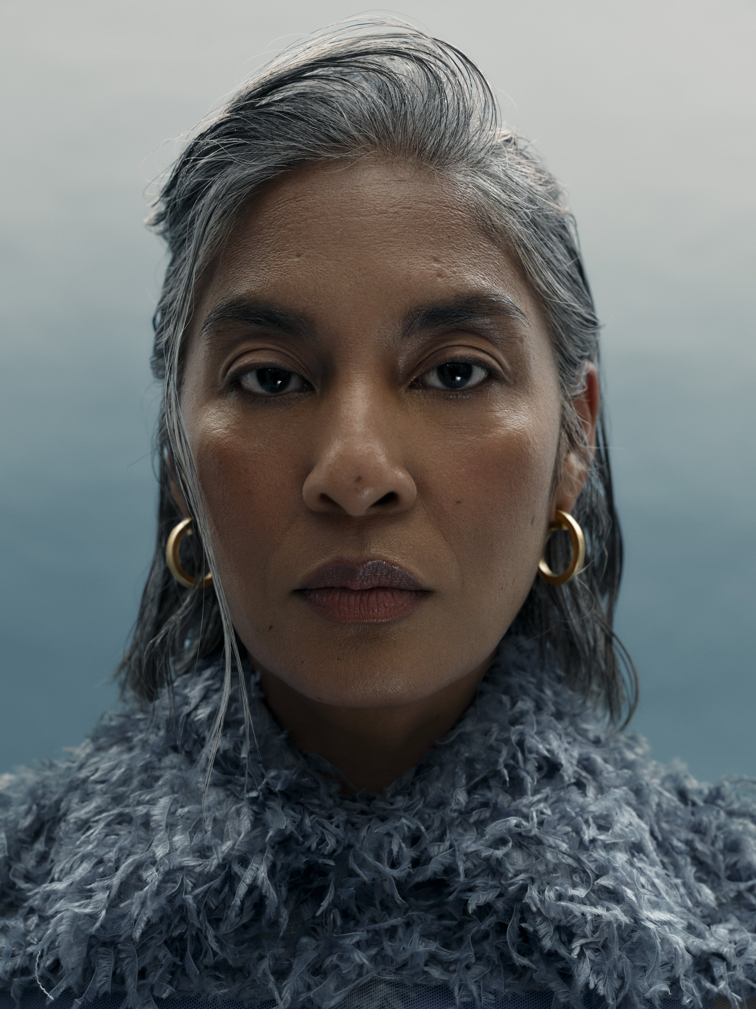Close-up portrait of a woman with shoulder-length gray hair, wearing gold hoop earrings and a textured, feather-like gray sweater, against a blurred, cloudy sky background.