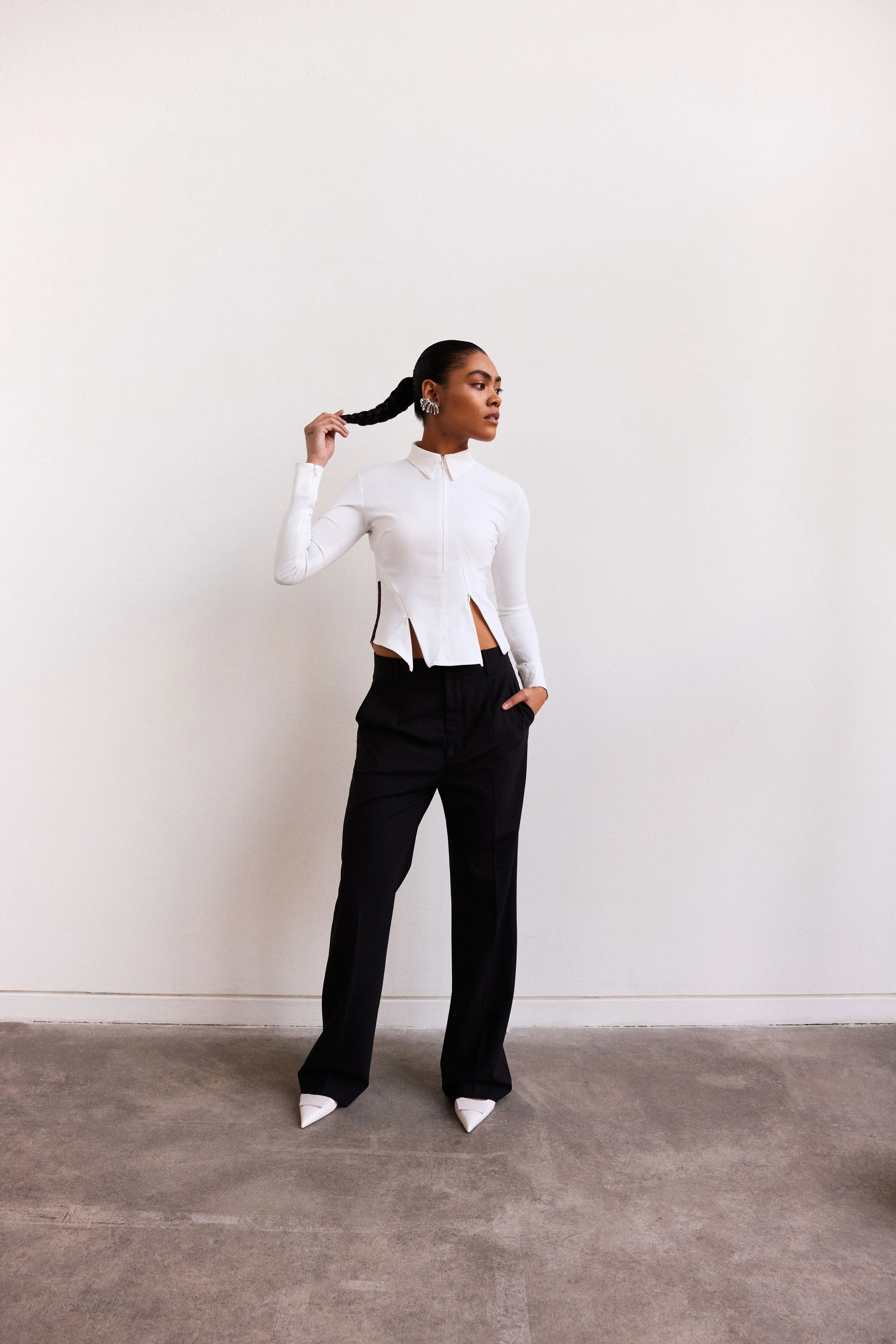 A woman with dark skin and long braided hair standing against a plain white wall, wearing a white zippers shirt, black pants, and white pointed shoes, with her left hand in her pocket and her right hand holding her braid.