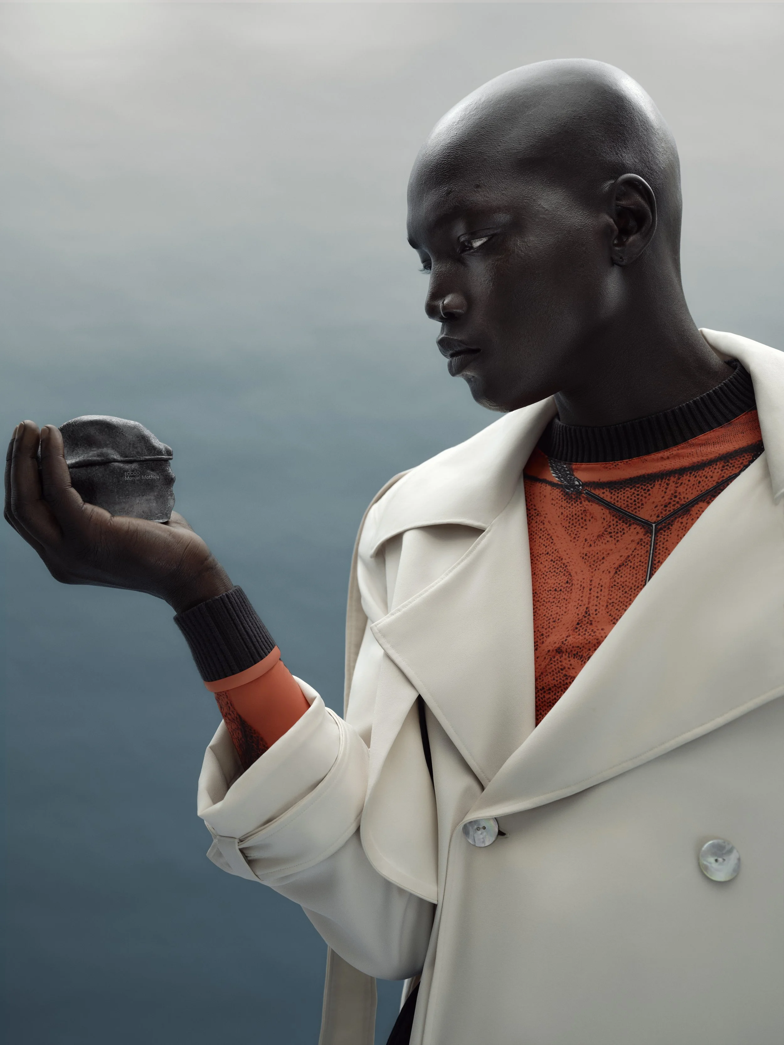 A person with dark skin, a bald head, wearing a white coat and black and orange clothing, is looking at a small black object in their hand against a cloudy sky background.