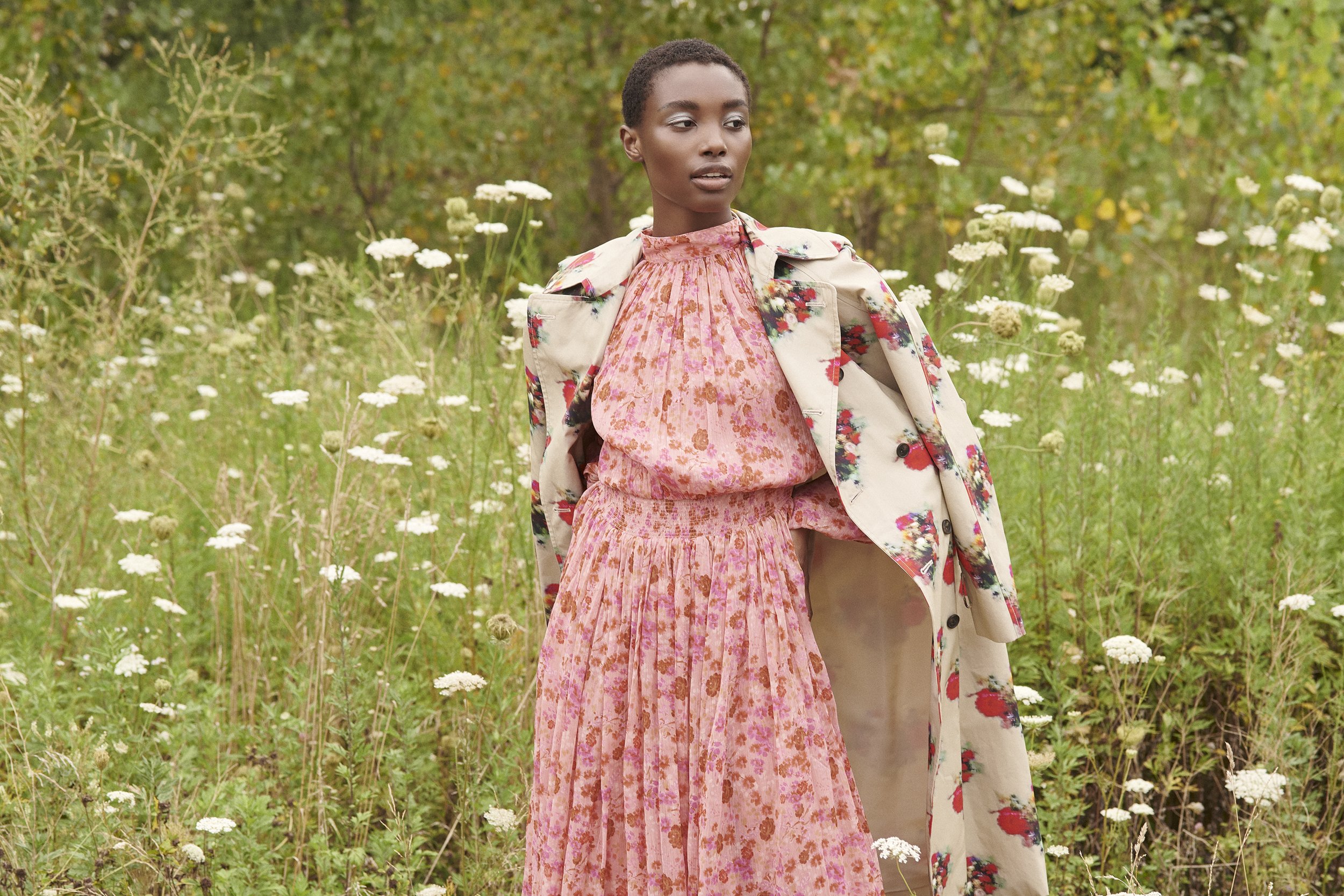 A woman in a floral coat and pink floral dress stands in a field of white flowers and greenery.