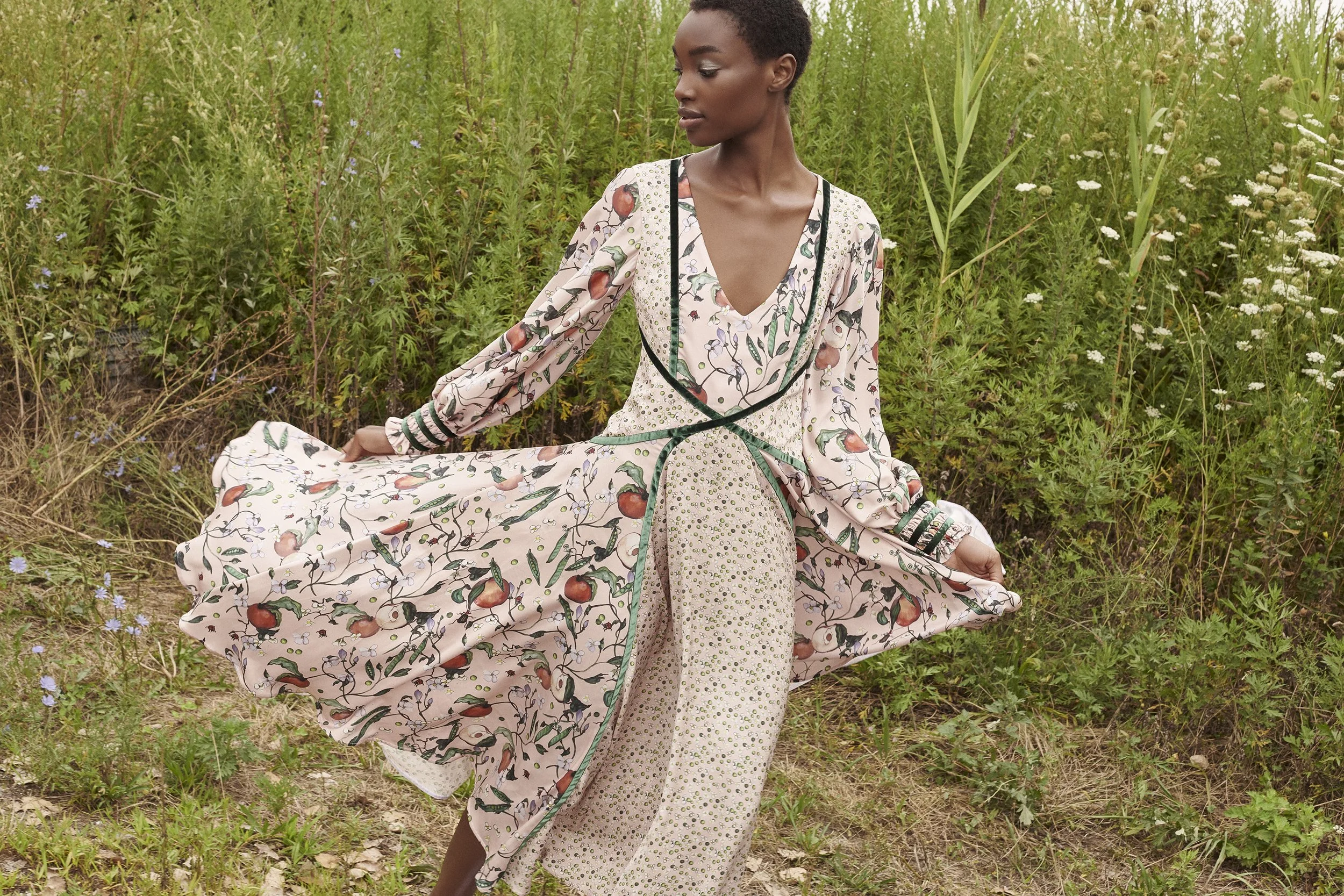 A woman in a floral maxi dress standing outdoors among green plants and wildflowers, holding the flowing skirt of her dress.