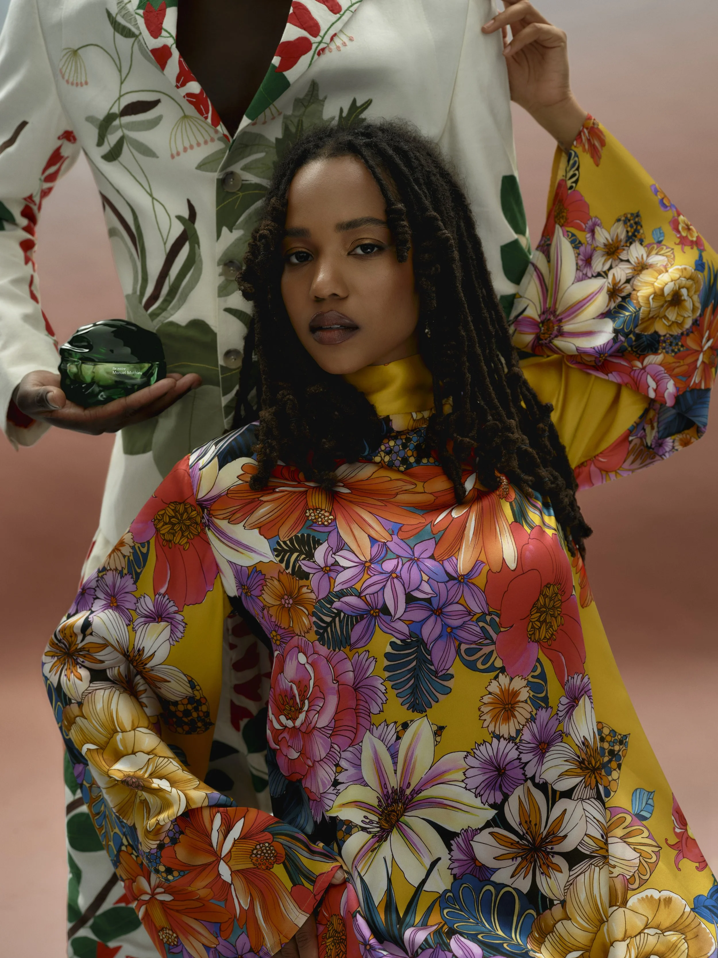 A woman with dark skin and long dreadlocks wearing a vibrant, multicolored floral dress, sitting with a confident expression, and a man standing behind her in a floral shirt, holding a green object in his hand.