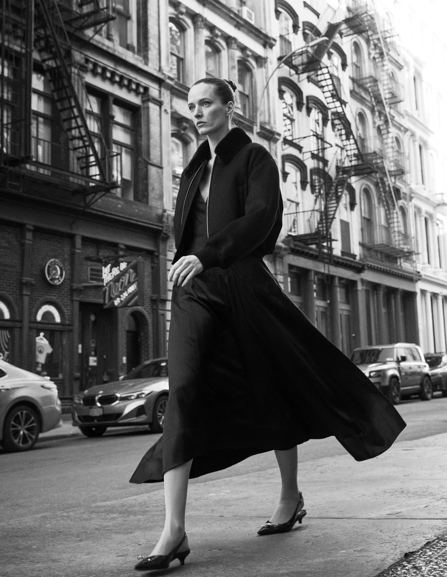 A woman walking across a city street in black and white. She is wearing a dark coat, a long flowing skirt, and high heels, with her hair tied back. The background shows a building with fire escapes and parked cars.