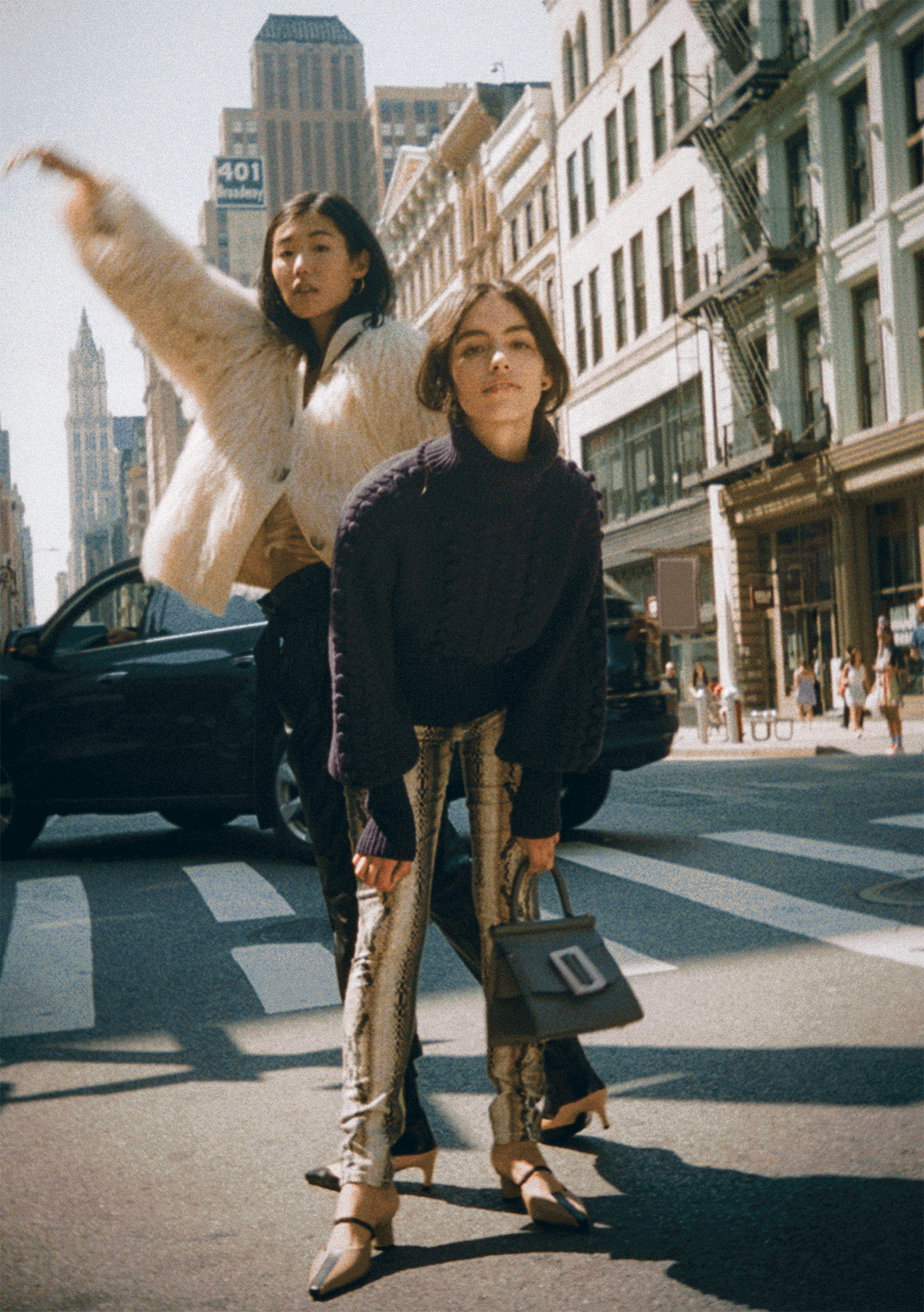Two fashionable women crossing a busy city street, with tall buildings and a clock tower in the background, dressed in trendy clothes and high heels.