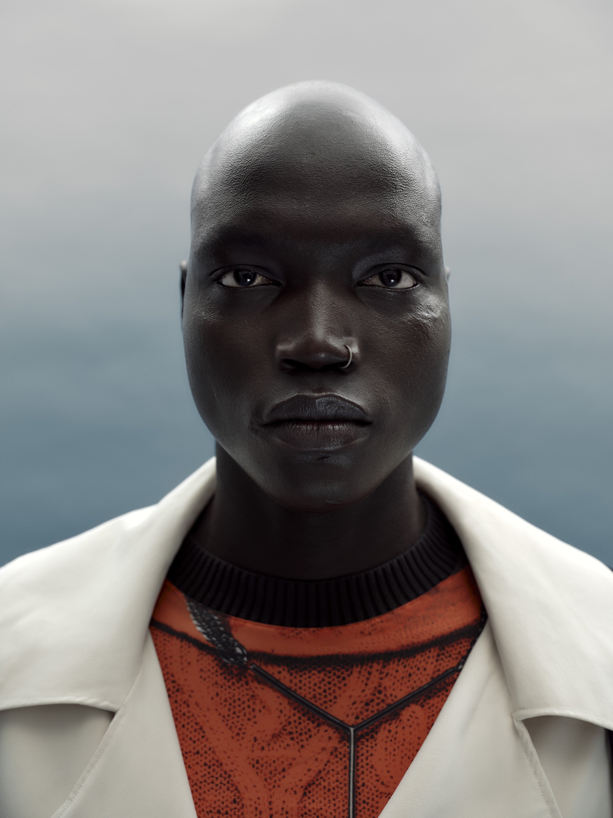 Close-up portrait of a person with dark skin, shaved head, wearing a white jacket and an orange patterned shirt, with a neutral background.