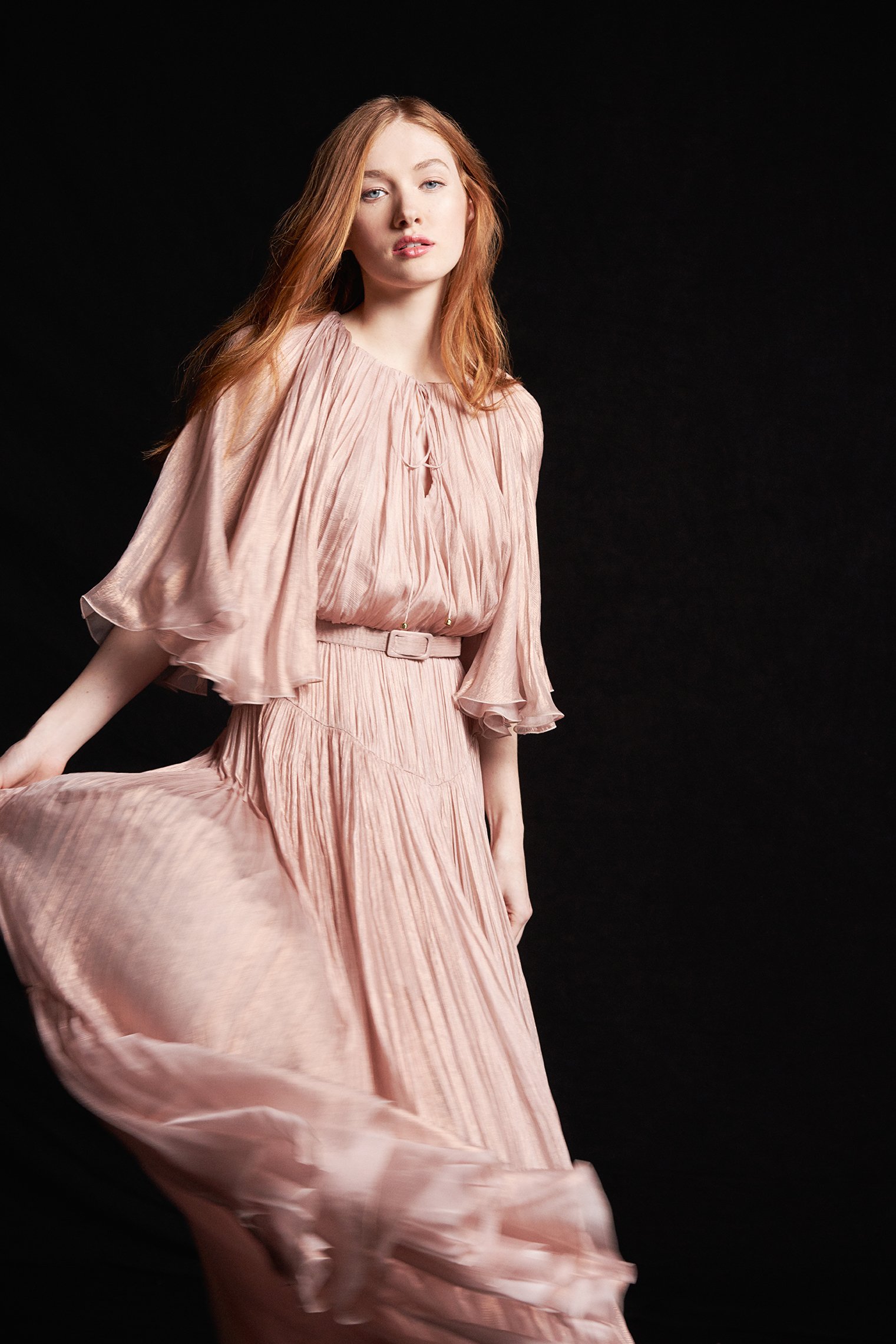 A young woman with red hair wearing a flowing pink dress against a black background.
