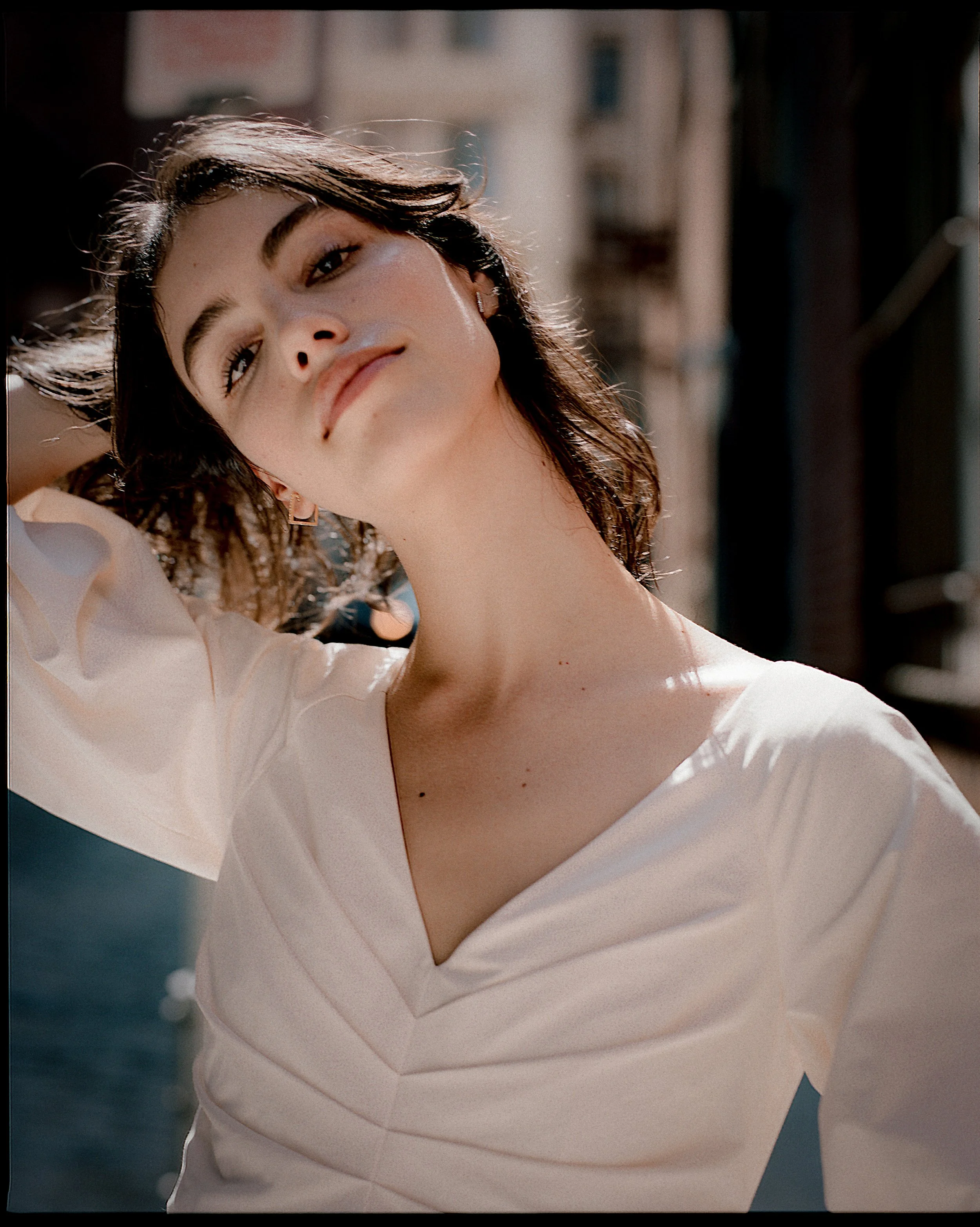 A young woman with dark wavy hair and light skin posing outdoors in sunlight, wearing a cream-colored V-neck top, with a background of buildings and a canal.