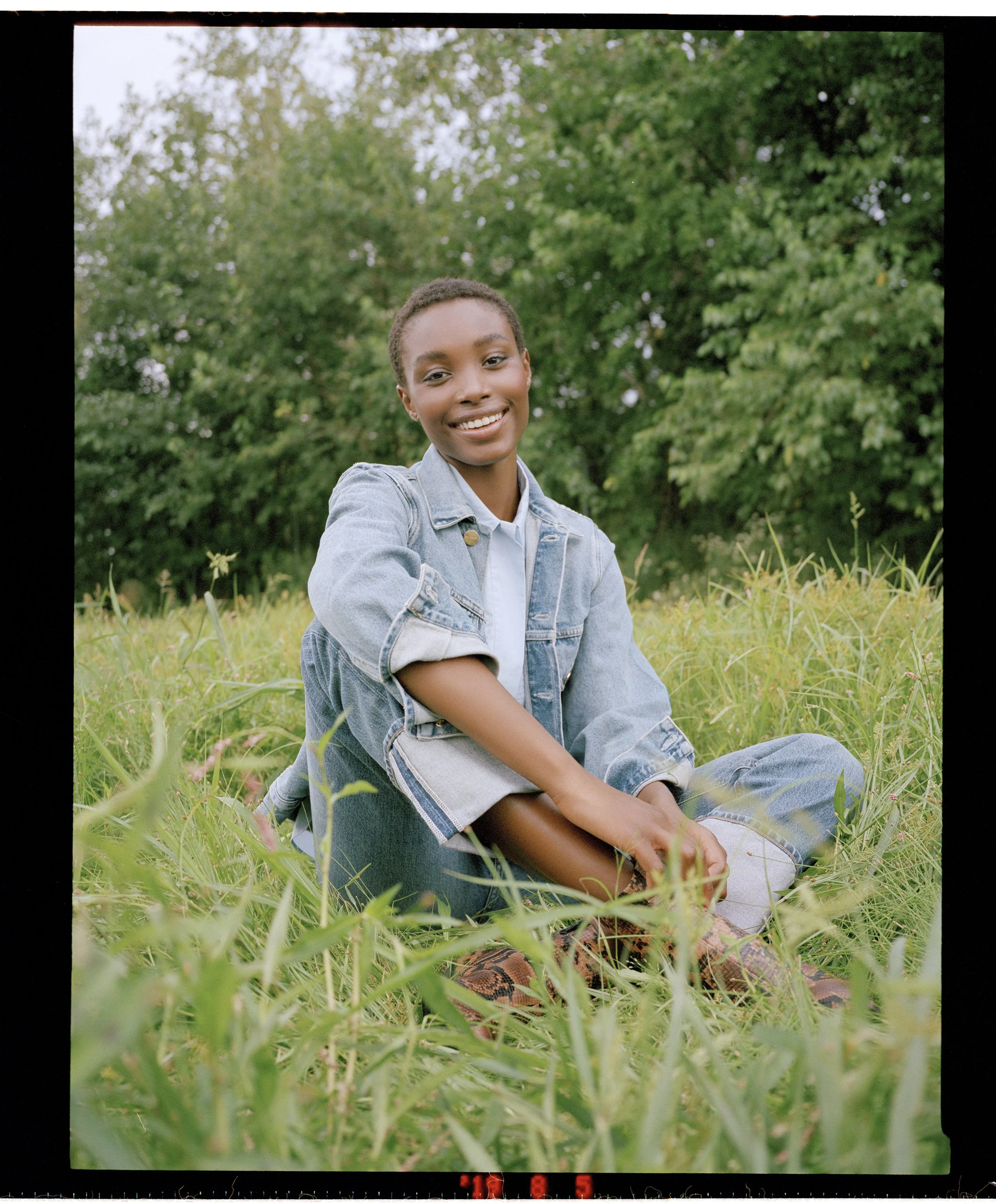 A smiling young woman sitting cross-legged in a grassy field with green trees in the background, wearing a denim jacket and jeans.