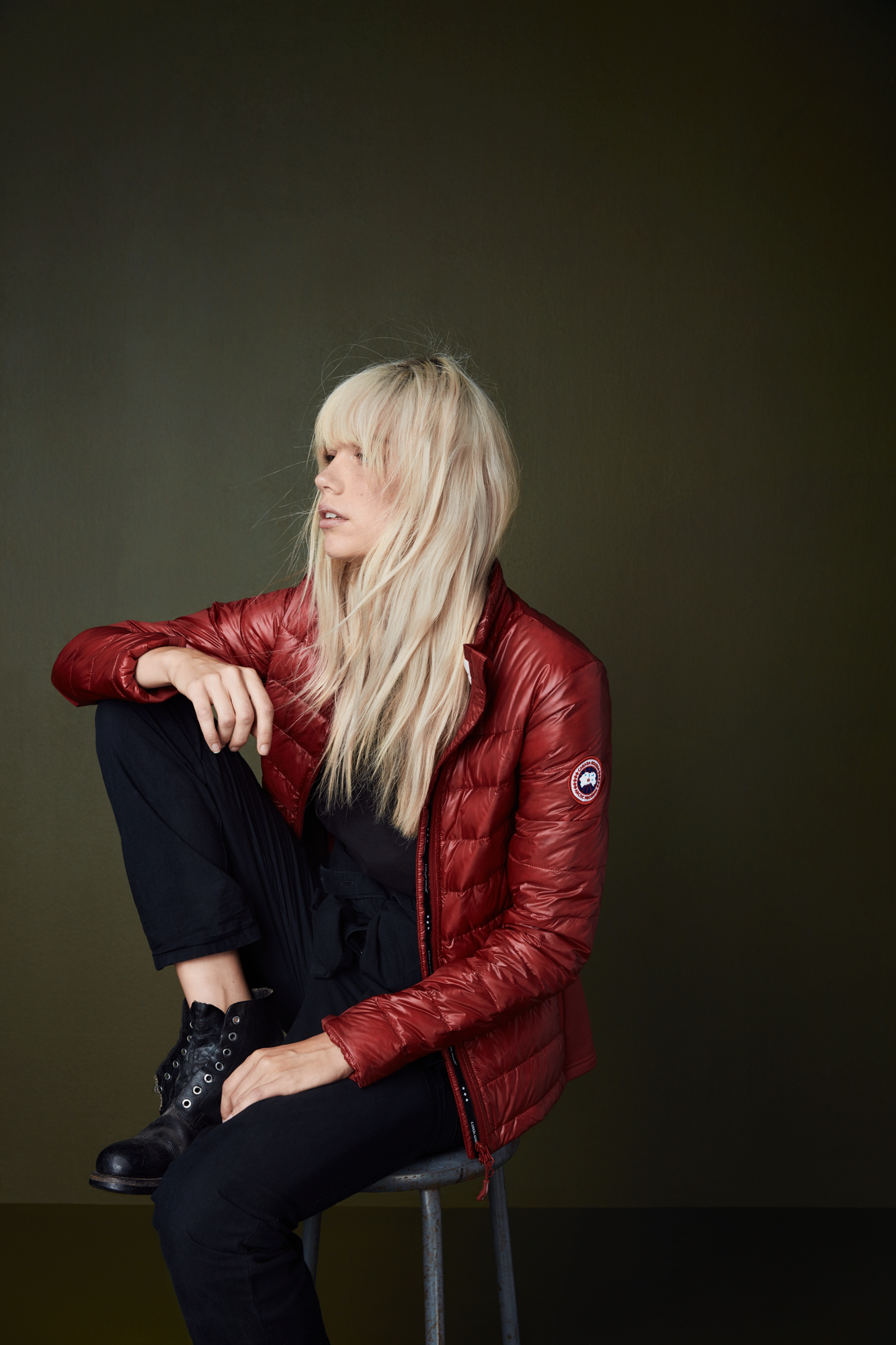A woman with long blonde hair, wearing a red jacket and black pants, sitting on a stool against a dark green background.