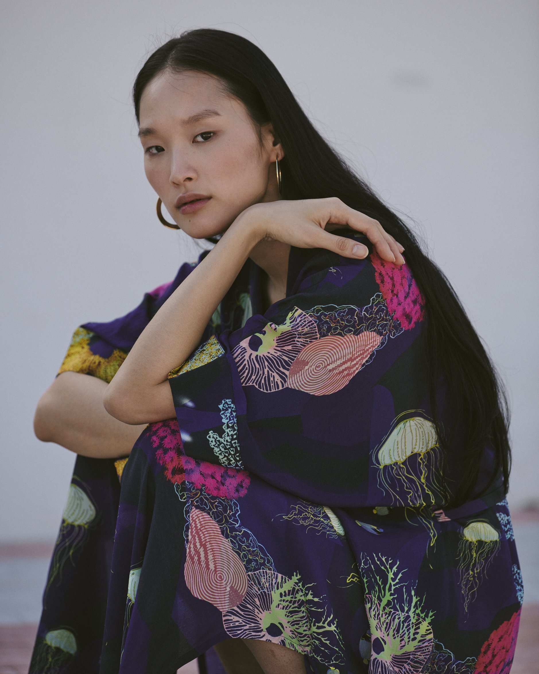 A woman with long black hair and gold hoop earrings, wearing a colorful floral-patterned dress, sits outdoors with a neutral background.