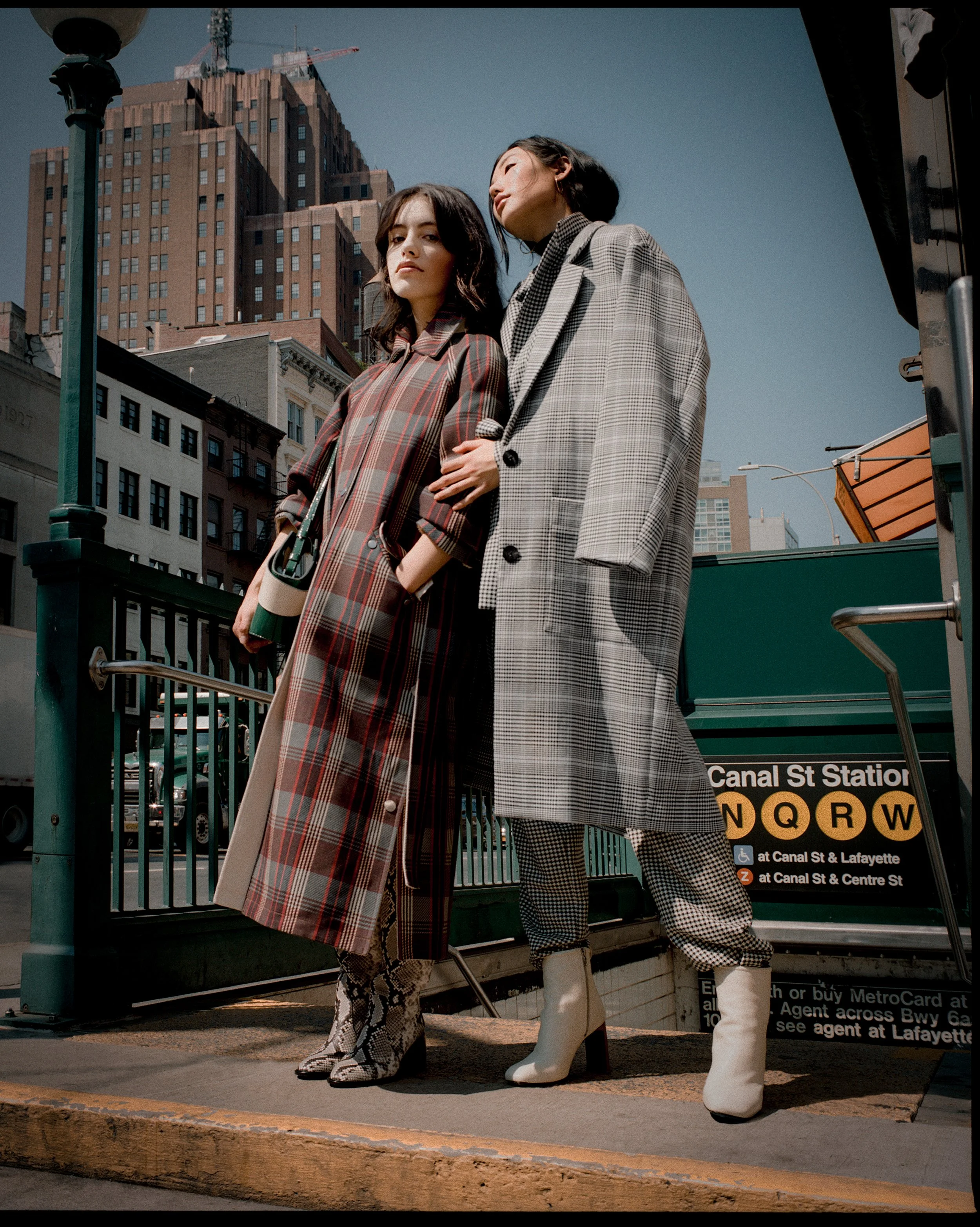Two women standing on a subway entrance in New York City, wearing fashionable plaid coats and boots, with tall buildings in the background.