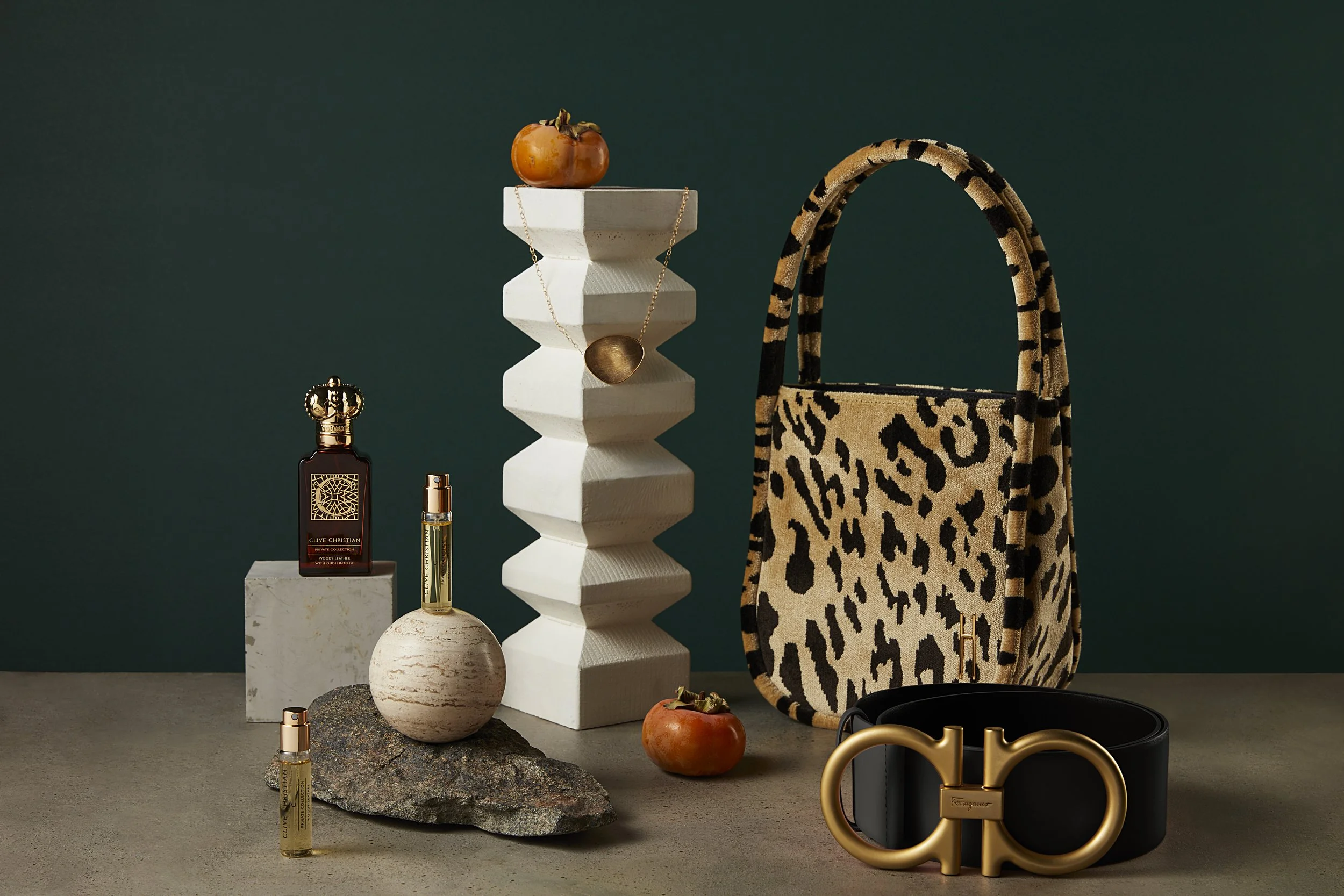 Display of luxury fashion items including a beige animal print handbag with black trim, a black belt with a gold buckle, a small dark perfume bottle, two small spray bottles, a white textured vase with a round fruit on top, a stone base with a spray 
