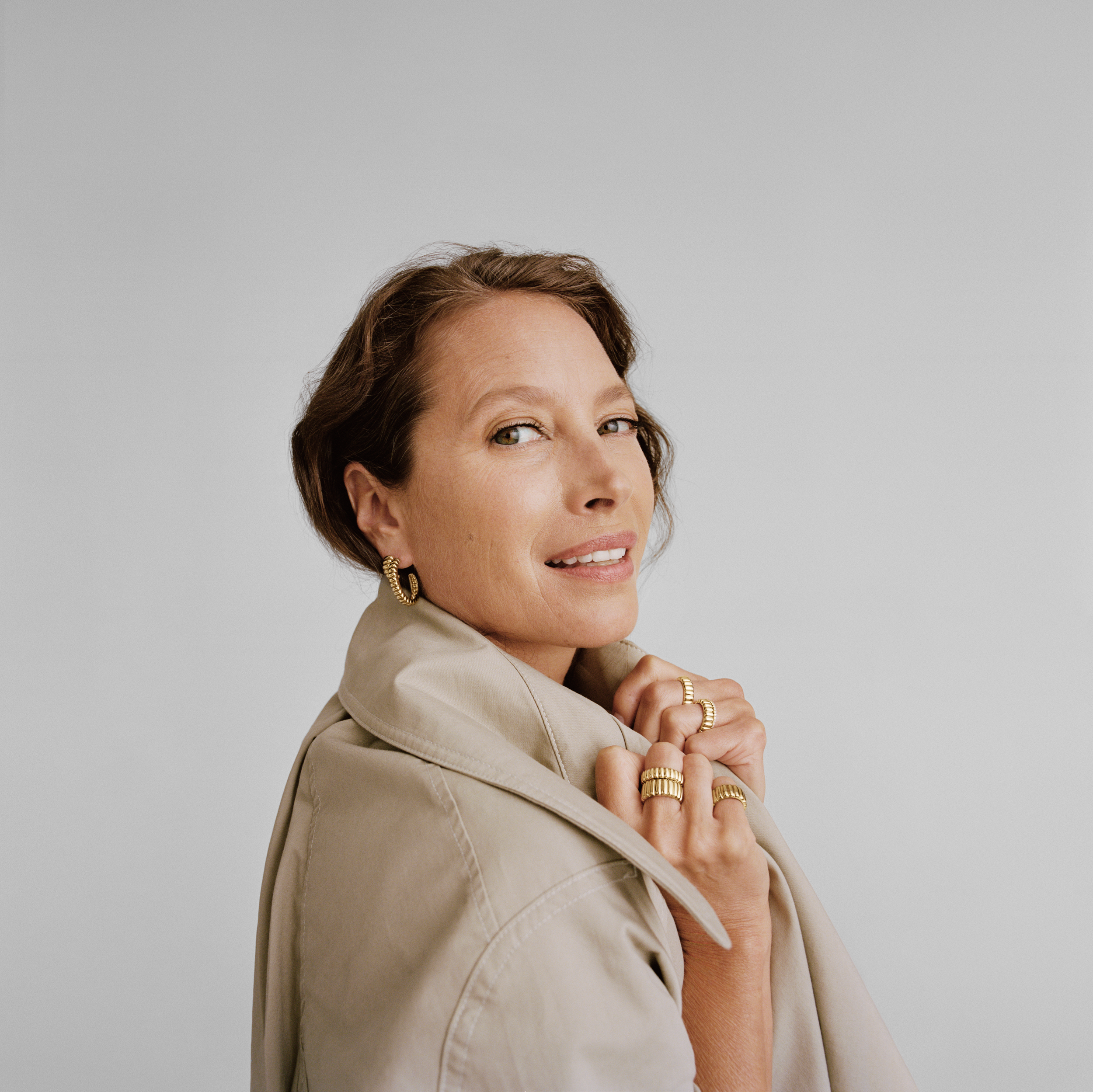 A woman with short brown hair, wearing gold jewelry and a light beige trench coat, poses against a plain light gray background.