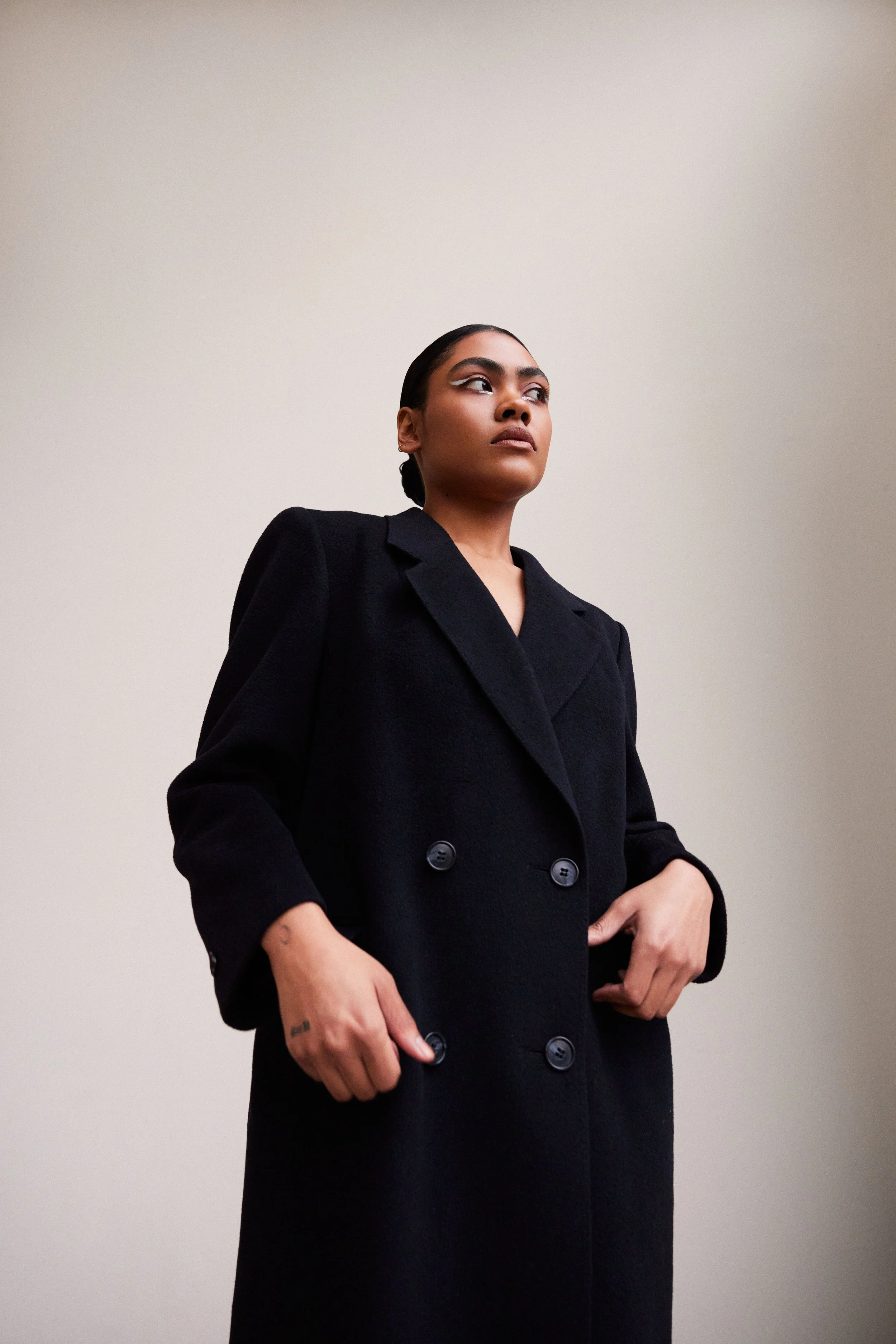 A woman with dark hair tied back, wearing a black coat, looking slightly upward, standing against a plain light-colored background.
