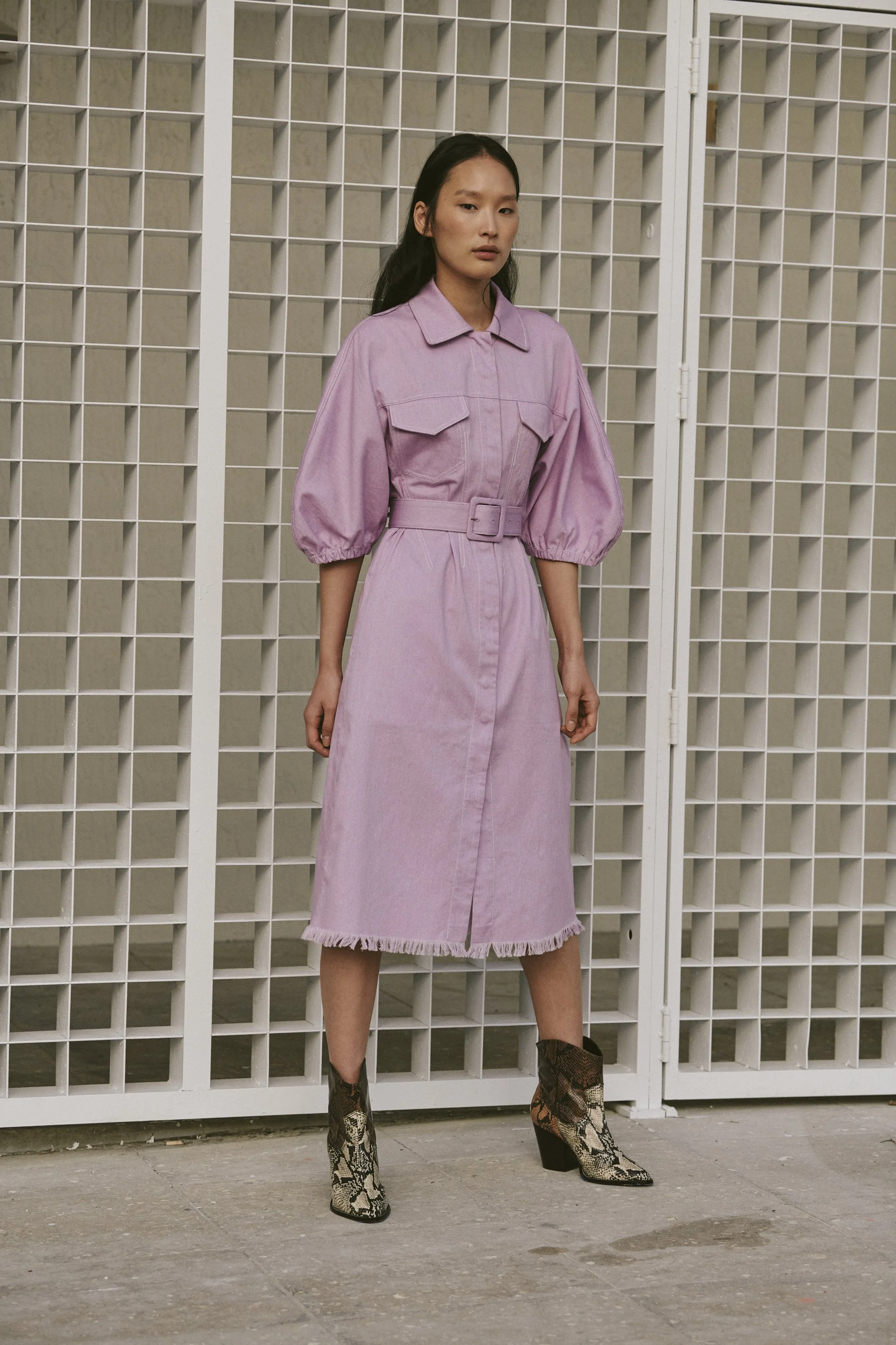A woman in a pink dress with puffed sleeves and a belt, standing in front of a white grid backdrop, wearing snakeskin ankle boots.