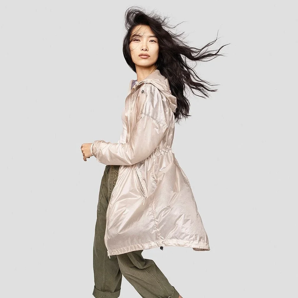 Woman with long black hair blowing in the wind, wearing a light-colored satin jacket and khaki pants, standing against a plain gray background.