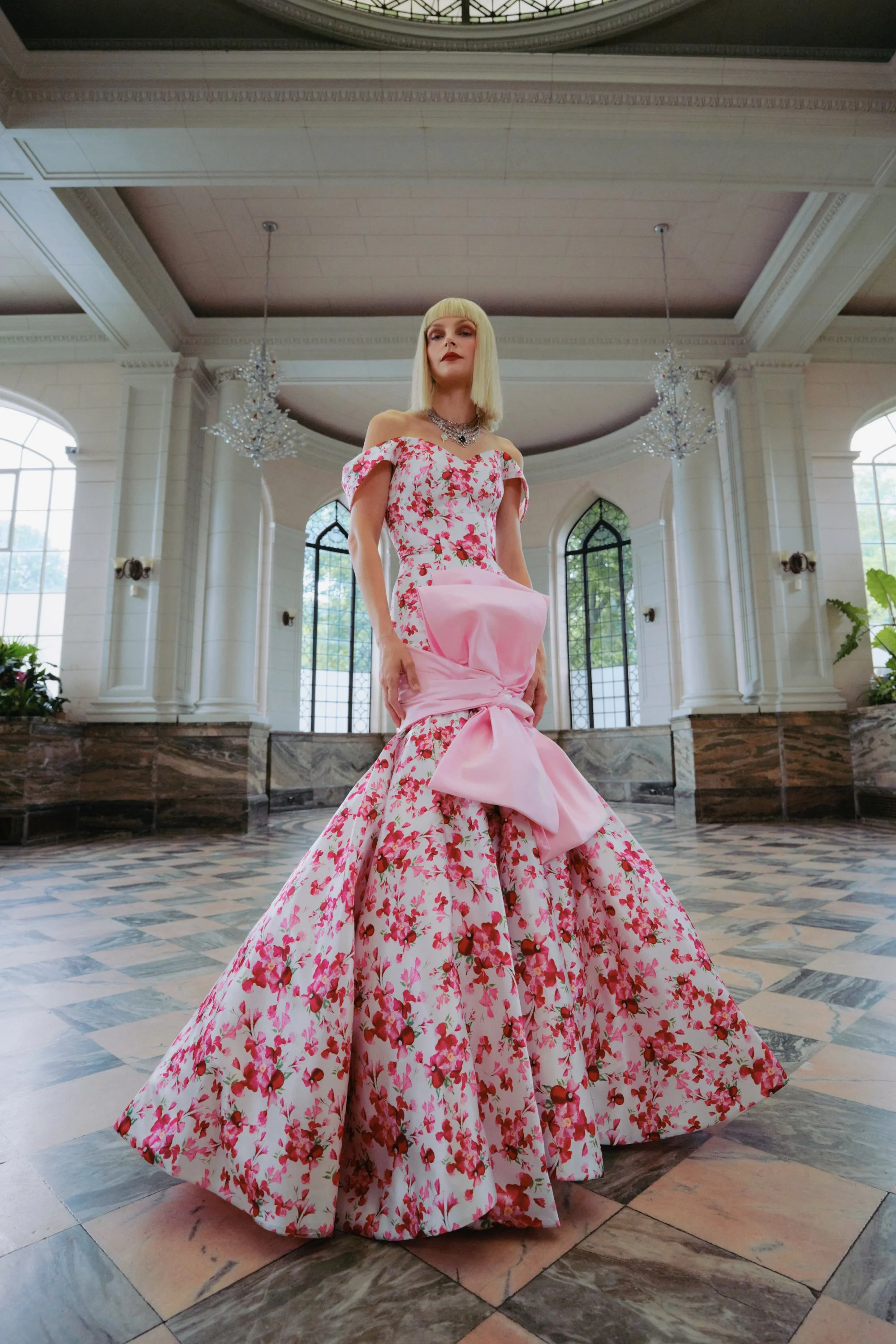A woman in an elaborate pink and floral gown with a large bow at the waist, standing in a grand, bright hall with tall windows, chandeliers, and marble flooring.