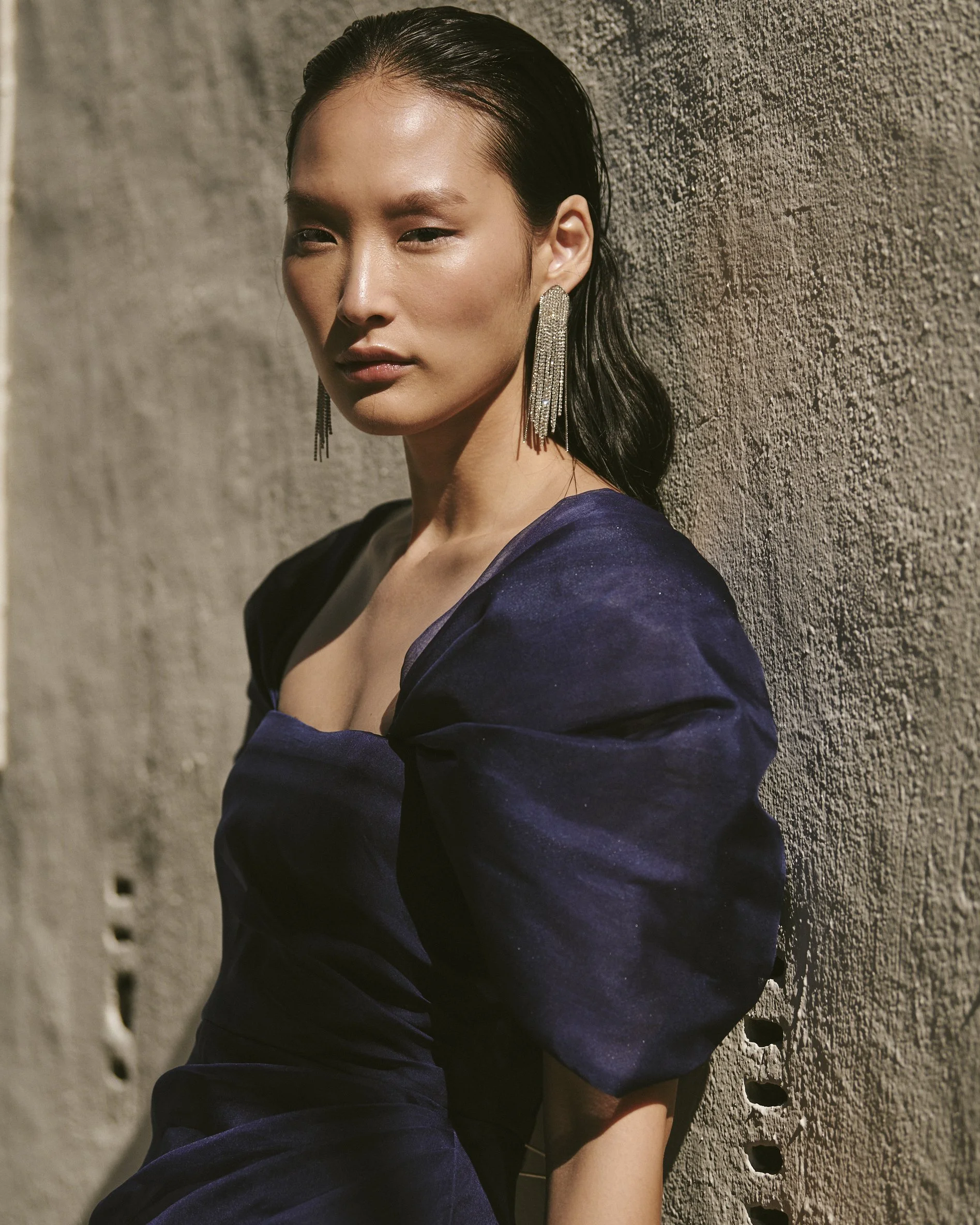 A woman with wet hair wearing a dark purple dress and large dangling earrings, posed against a textured concrete wall.