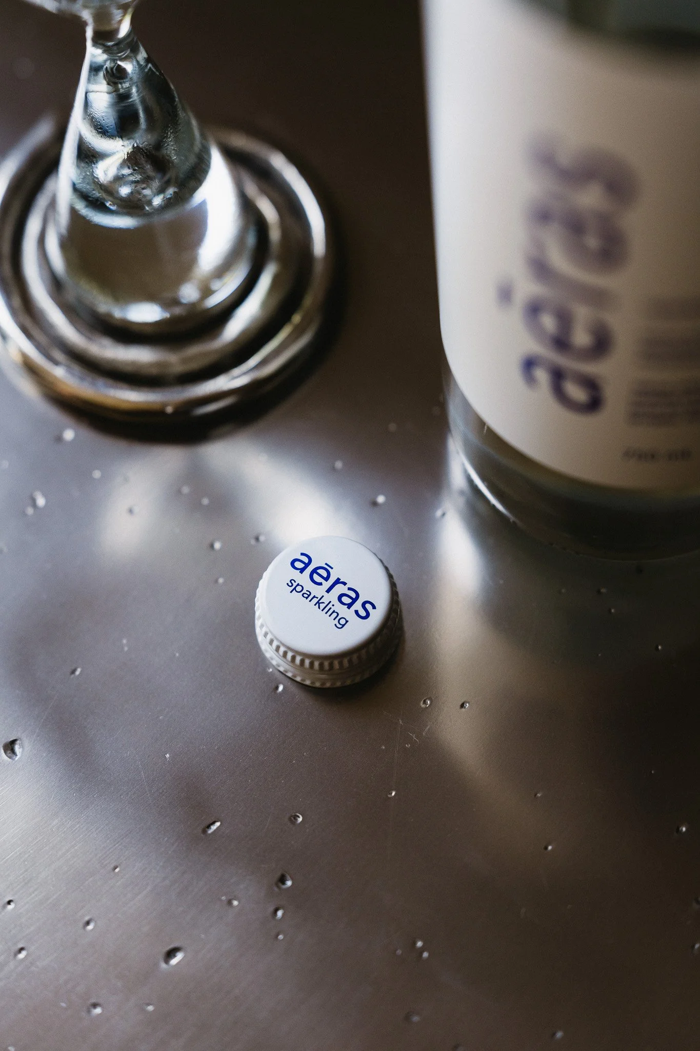 Close-up of soda bottle cap labeled 'aeras sparkling' on a wet metallic surface with a glass bottle and part of a white label in the background.