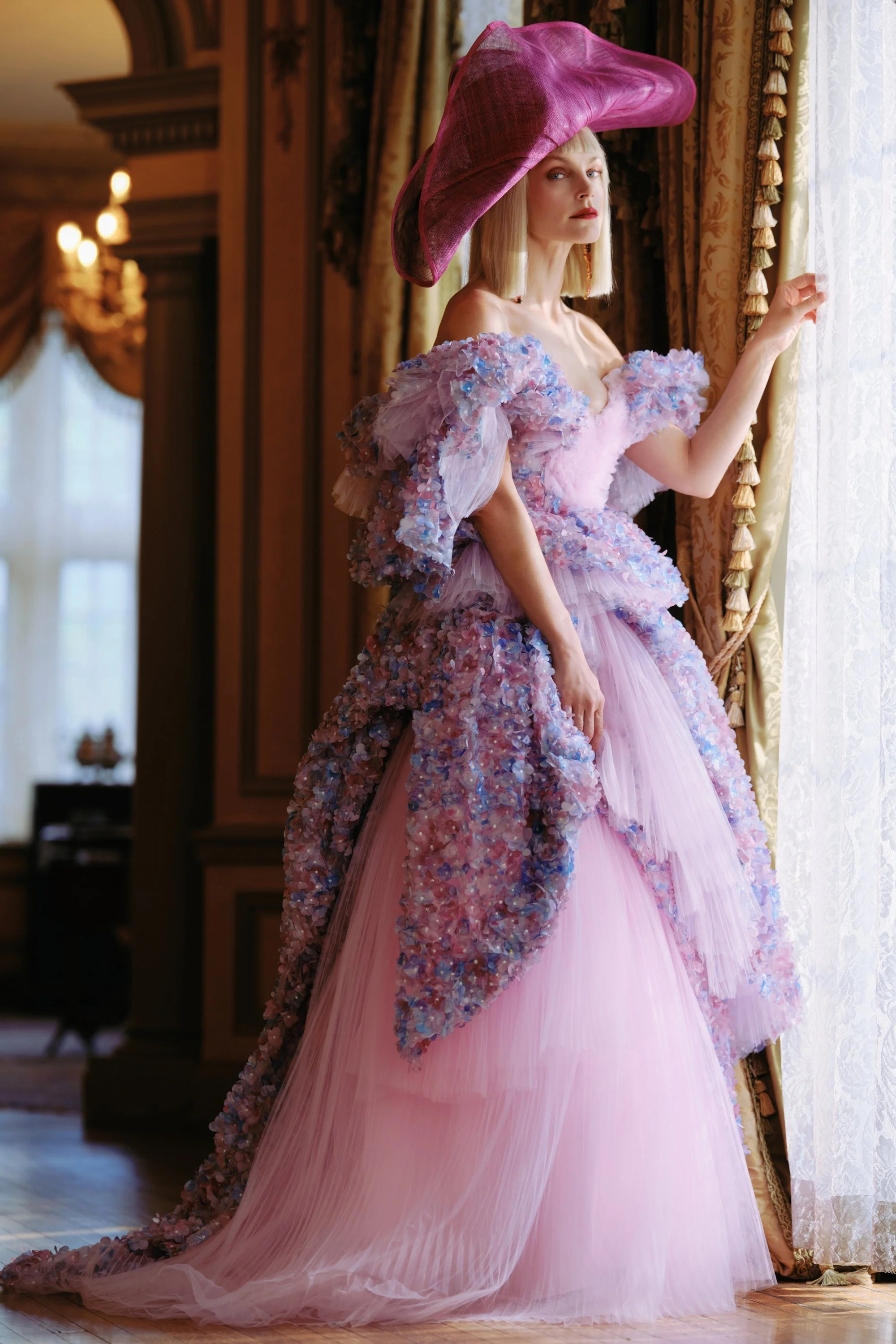 A woman in an elaborate pink and lavender gown with floral embellishments, standing by a window with golden curtains, wearing a large matching pink hat.