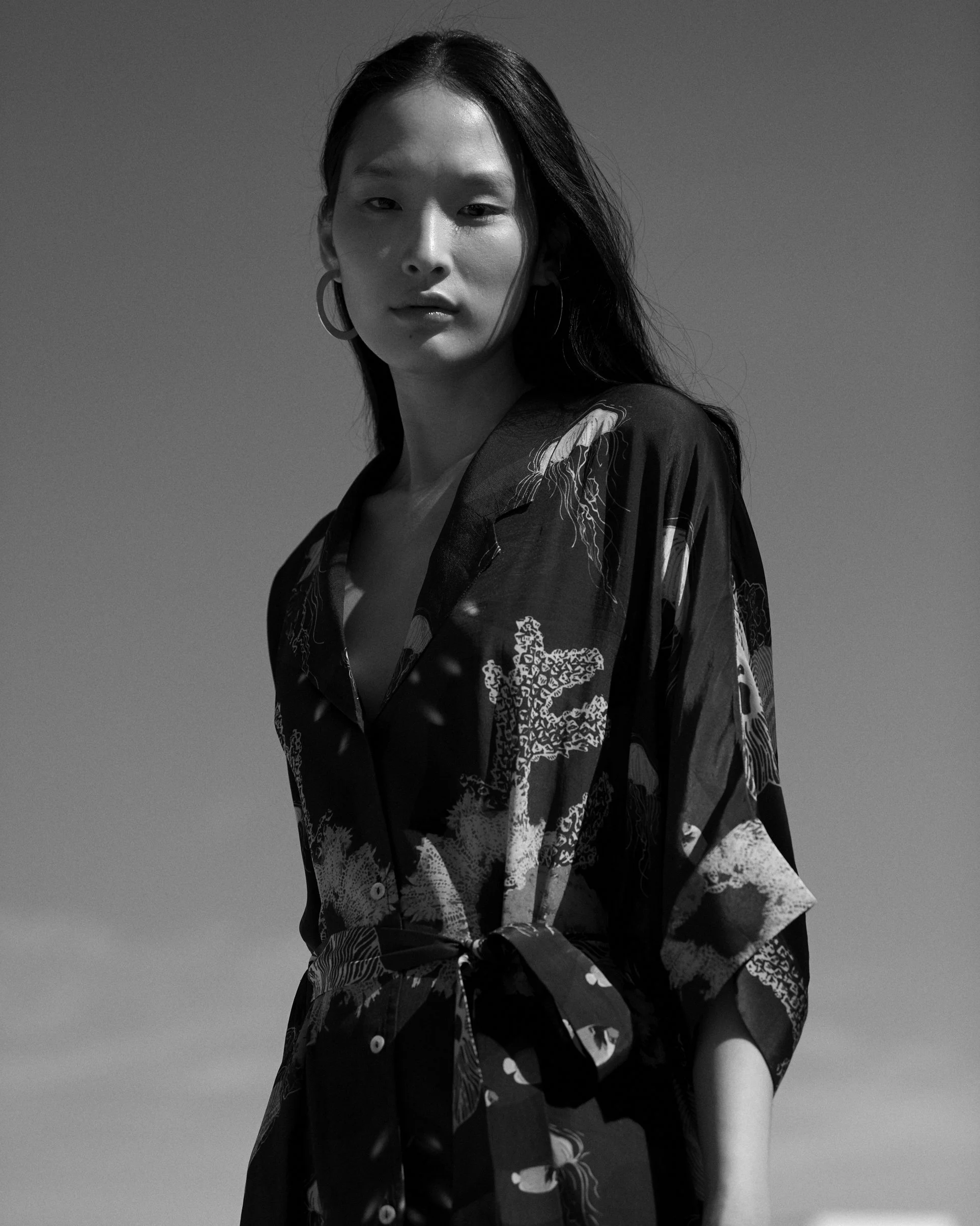 Black and white photo of a woman with long dark hair, wearing hoop earrings and a patterned blouse, standing outdoors against a clear sky.