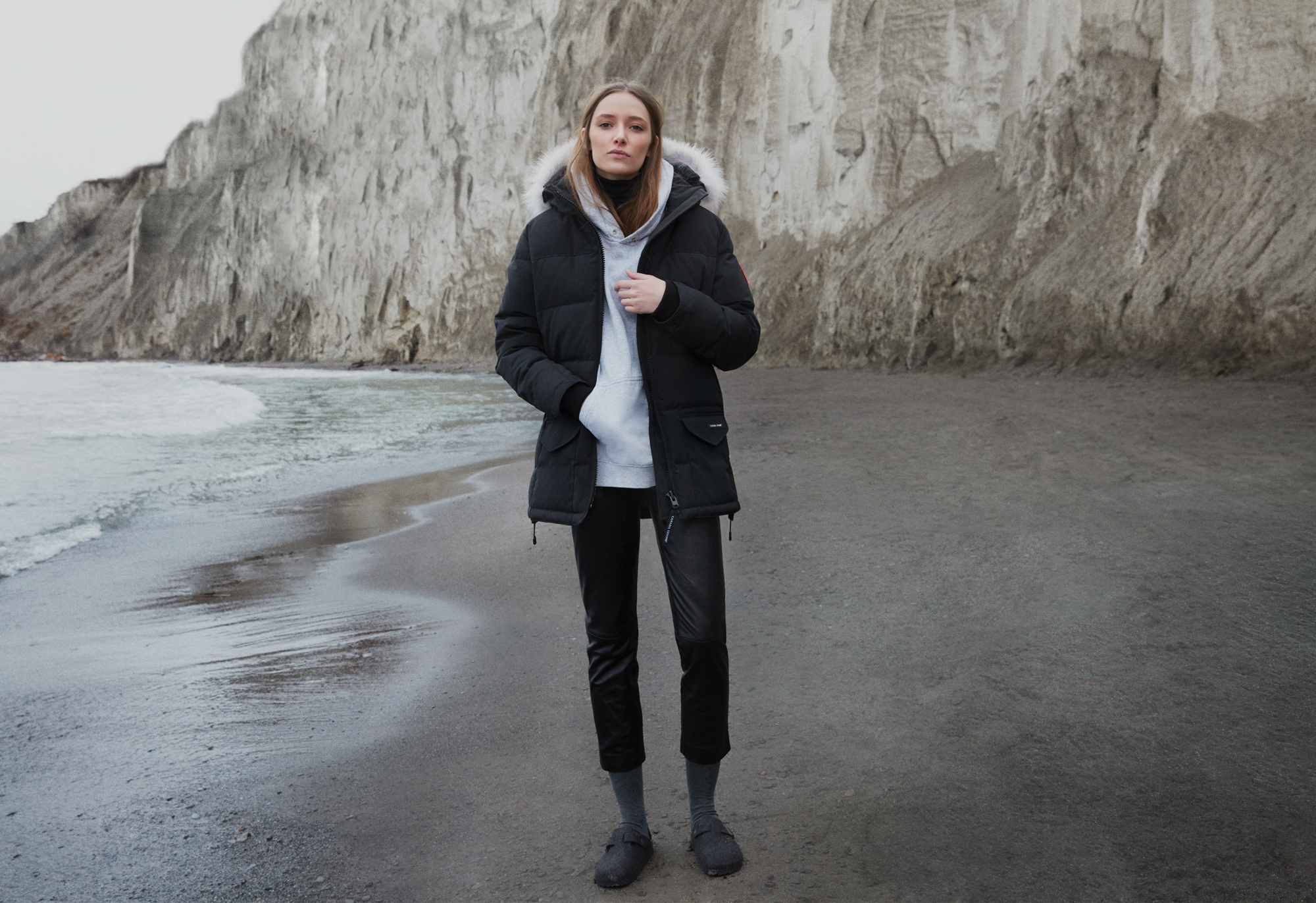 A woman in a black winter coat standing on a dark sandy beach with cliffs in the background.