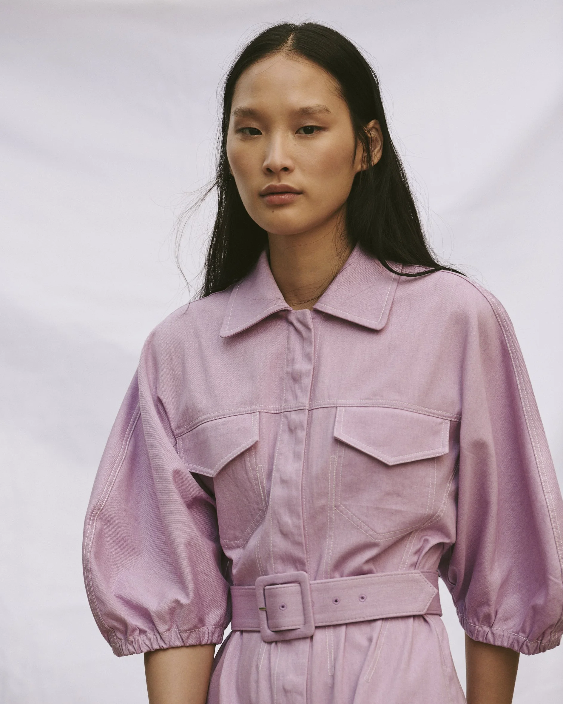 A woman with long dark hair wearing a pink jumpsuit with puffed sleeves and a belt, standing outdoors against a cloudy sky.