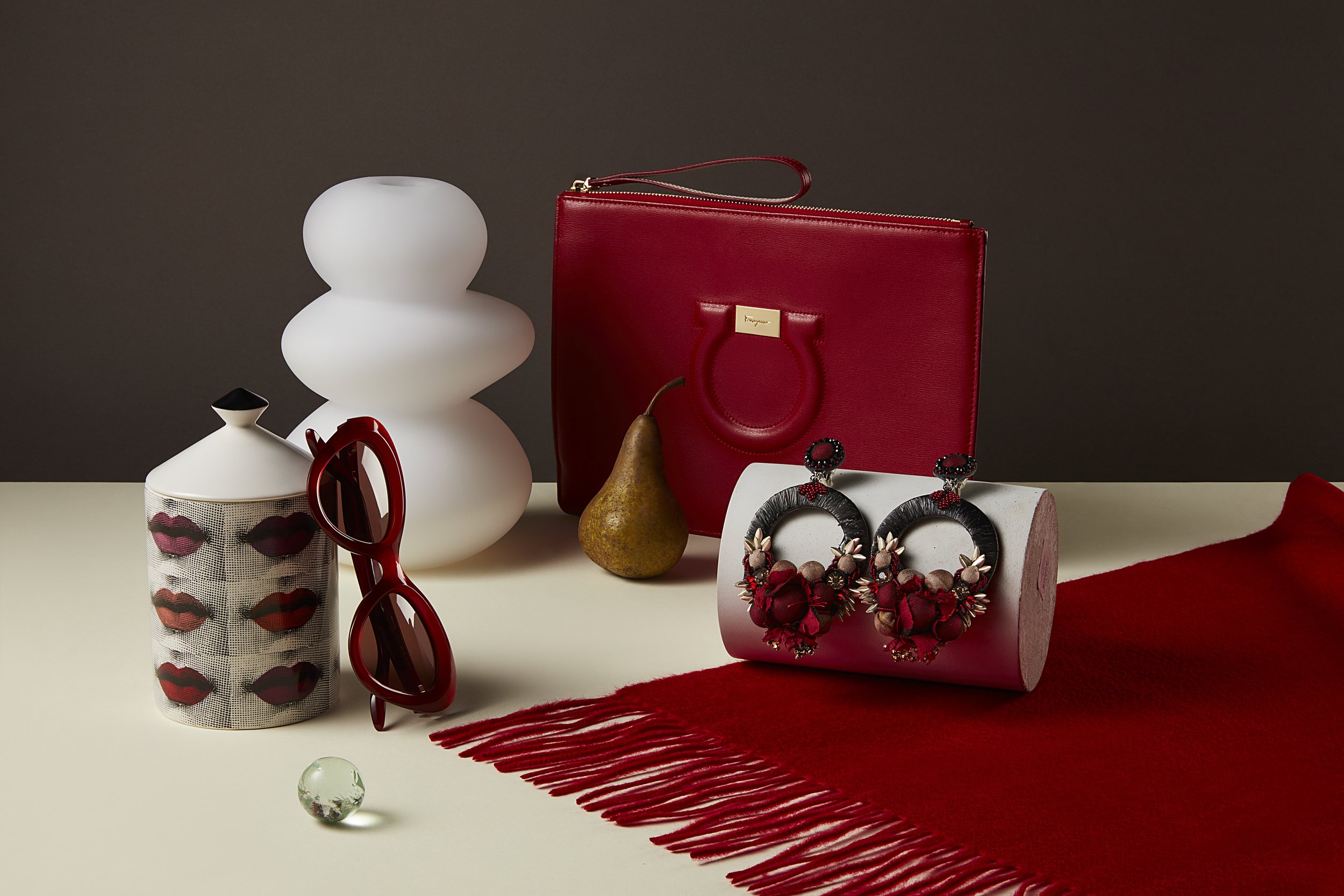 A still life arrangement featuring a white, abstract ceramic sculpture, red handbag, pair of red sunglasses, decorative jar with lips pattern, a pear, a red and black statement earrings display, a red fringed fabric, and a small glass marble, all set