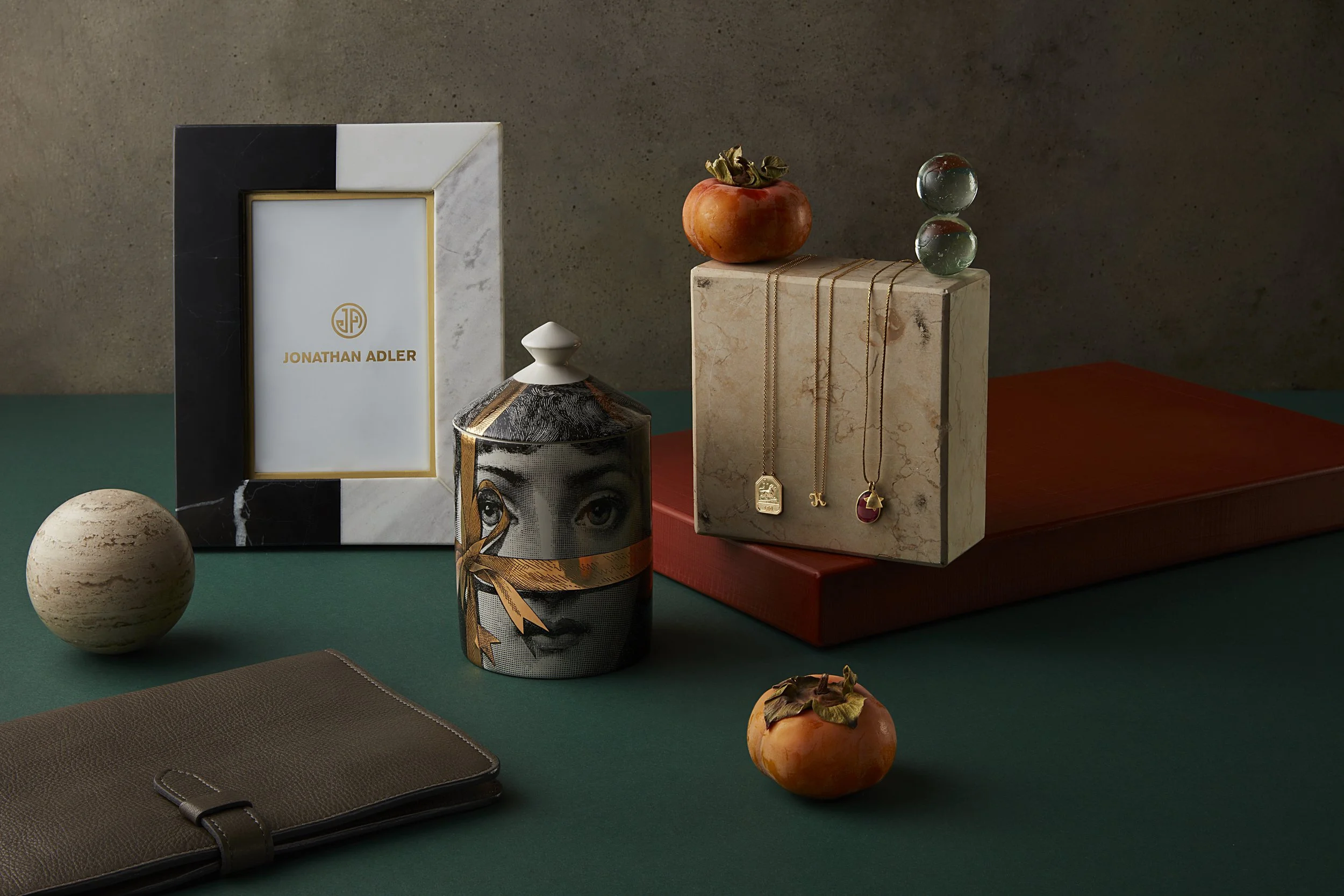 Still life arrangement with a Jonathan Adler photo frame, a jewelry box with necklaces, a cylindrical container with an eye and face design, a small beige sphere, a leather notebook, a pomegranate, and a flat red item on a green surface against a gra