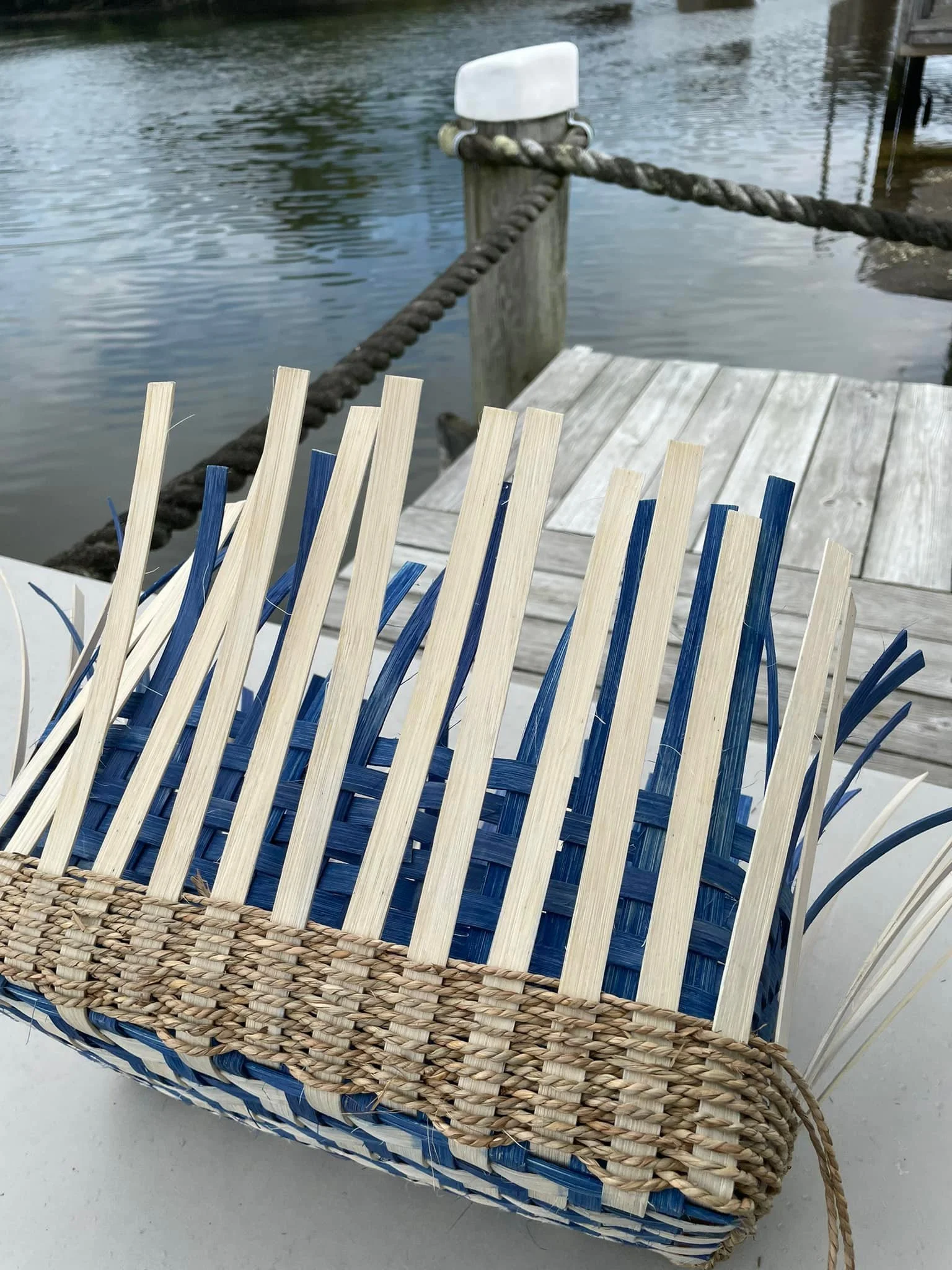 A woven basket with a blue and natural straw pattern, placed on a white surface near a dock by the water