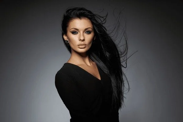 Portrait of a woman with long black hair wearing a black top, posing against a gradient gray background.